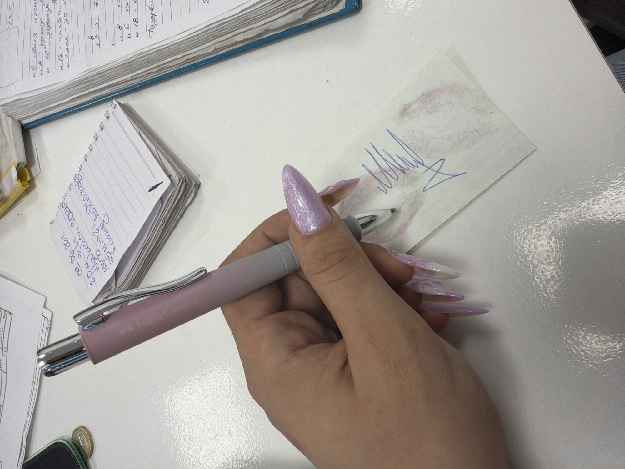 The image depicts a close-up view of a person's hand with long, acrylic nails holding a pink and silver Faber-Castell pen. The pen is being used to write on a small, torn piece of paper that has a faint pink stain, possibly from lipstick, and a blue ink signature or scribble. The background shows a white desk surface with various papers, including a notepad and a bound ledger. There are no discernible landmarks or contextual clues to suggest a specific location within Stara Zagora, Bulgaria. The scene appears to be an indoor workspace or desk area.
