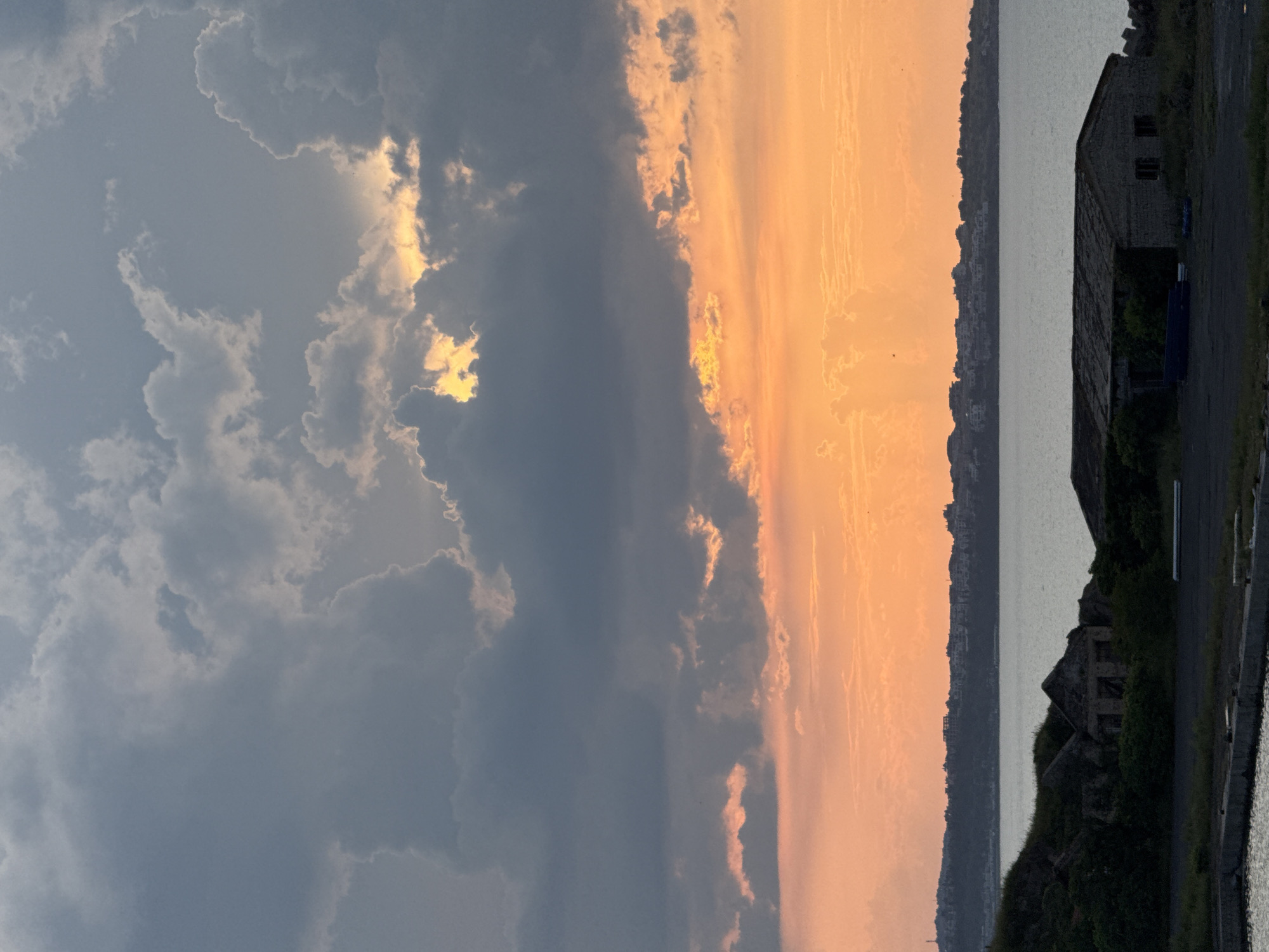 The image presents a dramatic coastal landscape captured during what appears to be sunset in Sozopol, Bulgaria. The sky is a prominent feature, strikingly divided into two distinct atmospheric conditions. On the left side, dark, heavy clouds in shades of gray and deep blue dominate, with bright, golden sunlight piercing through gaps, illuminating their edges and creating strong highlights. This contrasts sharply with the right side of the sky, which is awash in warm hues of orange, peach, and soft yellow, indicating the presence of the setting sun.
Below the expressive sky lies a calm body of water, likely the Black Sea, which reflects the subdued light. A distant landmass or coastline stretches across the horizon, appearing as a dark silhouette against the vibrant sky, with subtle hints of urban development or buildings along its ridge. In the immediate foreground on the right, a dark, weathered stone building, possibly an old structure or ruin, is partially obscured by dense, dark foliage. An unpaved or paved area is visible next to the building, leading towards the water.
No people are visible, and no specific activities are occurring beyond the natural progression of the evening. The scene highlights a moment of intense natural beauty and stark contrast between light and shadow, suggesting powerful weather patterns alongside the serene presence of the sea and land. There is no visible text in the image.