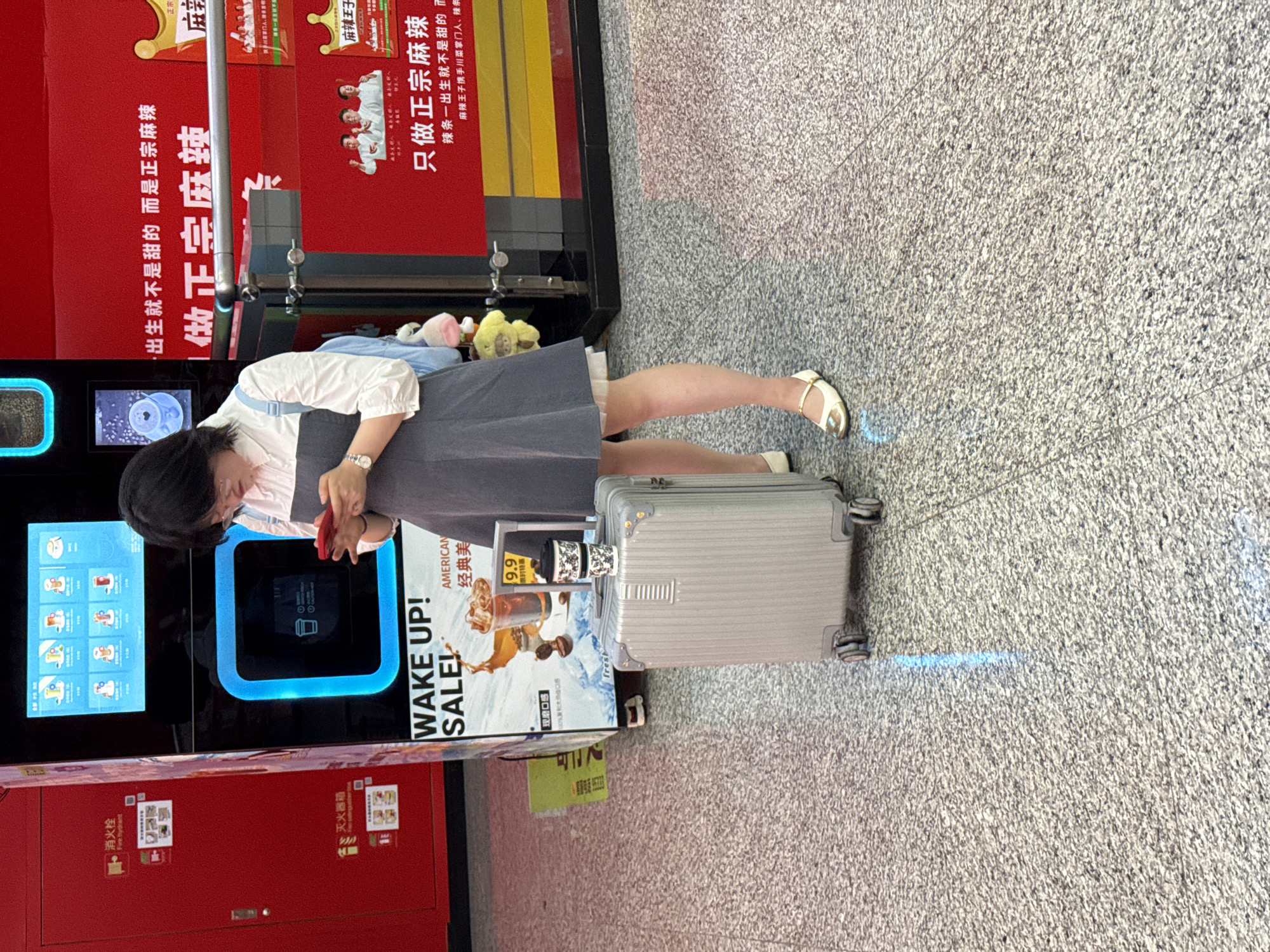 A woman wearing a white top and grey dress stands next to a vending machine in Yuzhong District, China. She appears to be interacting with the machine using her phone. A silver suitcase sits next to her. The vending machine has various drink options displayed on the screen. Adjacent to the machine, a red panel has fire extinguisher instructions and "Fire Hydrant" written in Chinese characters. Behind the woman and the vending machine are advertisements, including one for "Ma La Wang Zi," a type of spicy snack. The floor is tiled in gray. An advertisement next to the vending machine has an image of coffee and text, "Wake Up! Sale! American Classic Beauty."