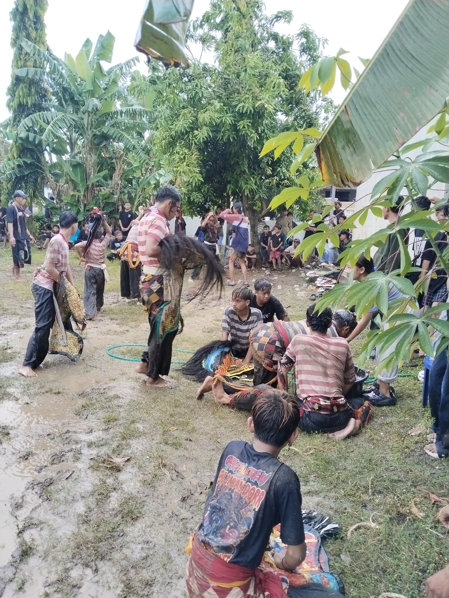 The image depicts a muddy outdoor scene in Karang Tengah, Indonesia, where a group of people are participating in a traditional ritual or performance. The ground is wet and covered in mud, suggesting recent rain.

Several individuals are dressed in striped shirts and traditional attire, possibly preparing or cleaning decorative horse masks. One man stands holding a mask with a long black mane. Others are sitting on the ground, seemingly working on the masks or resting. In the background, a larger group of onlookers observes the event. Trees and foliage surround the area.