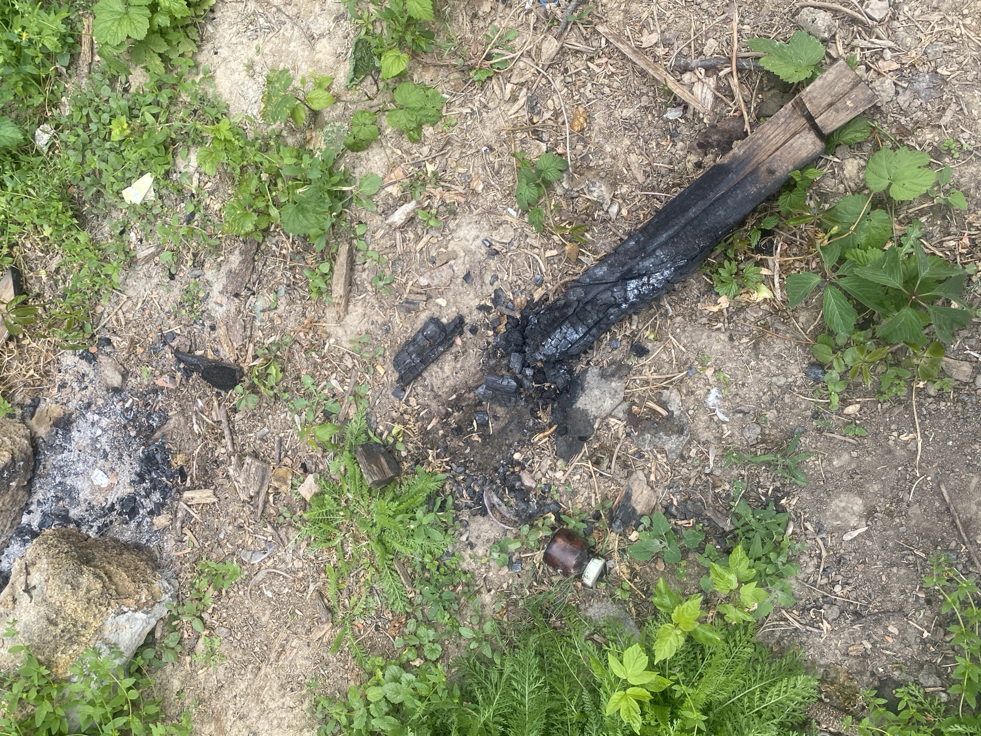 The scene depicts a patch of bare ground in Matiashivka, Ukraine, interspersed with green vegetation. Central to the scene is a partially burned wooden beam resting on the ground, surrounded by charred remnants of a fire. A small, dark-colored bottle lies among the ashes. A pile of ash and stones, suggestive of a former fire pit, is located on the left side of the frame. The ground consists primarily of dirt and scattered debris. No people or animals are visible.