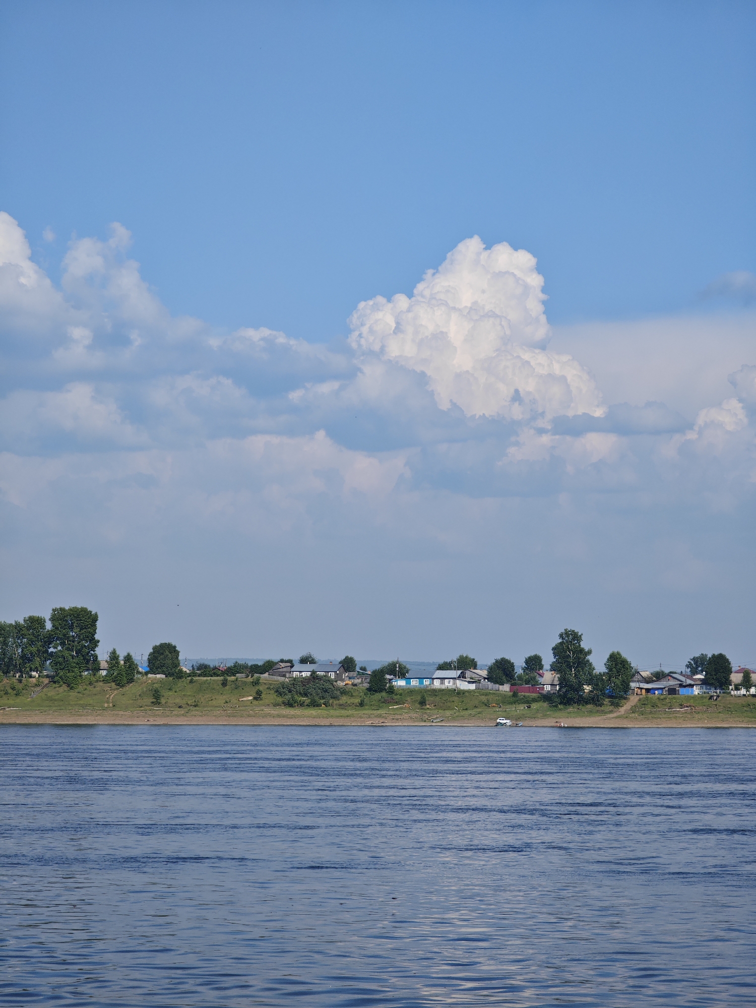 This image depicts a wide, blue river with gentle ripples under a bright blue sky filled with fluffy white clouds. Along the distant riverbank, a small village or settlement is visible, characterized by a line of houses, some with blue and red roofs, nestled amongst green trees. The houses appear to be of a traditional, rural style. The landscape is verdant, with the riverbank covered in grass and dotted with trees of various sizes. A small white vehicle is parked on the bank near the water. The scene is serene and suggestive of a peaceful, rural environment in Russia.