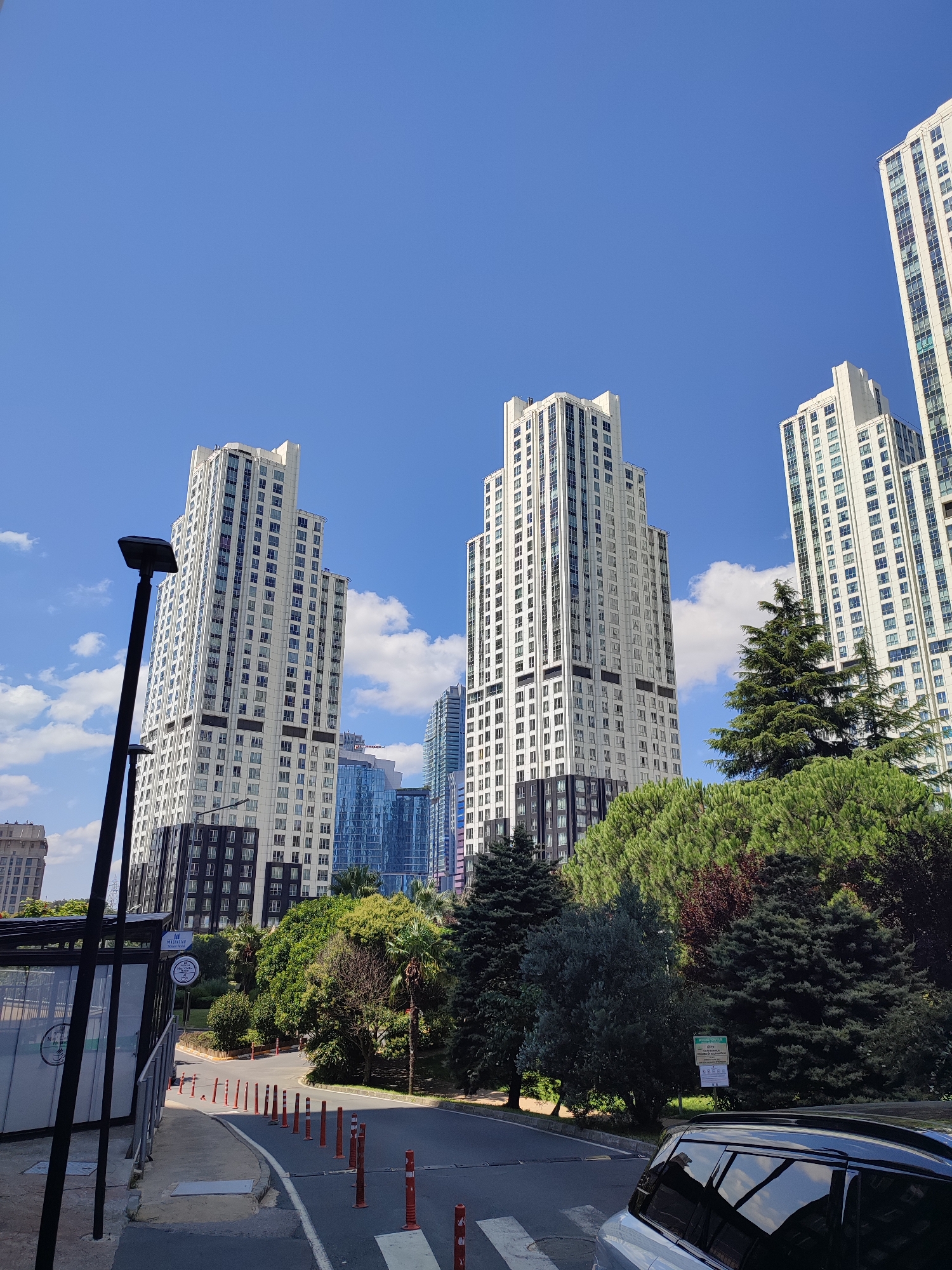The image shows a modern cityscape with several tall, white buildings under a clear blue sky with scattered clouds. In the foreground, a paved road with orange traffic cones and a pedestrian crossing is visible. To the left, a bus stop shelter with a black pole and signage is present. A black SUV is parked on the right side of the road, reflecting the surrounding environment. The buildings are characterized by their numerous windows and dark decorative bands. Lush green trees and foliage surround the base of the buildings, suggesting a park or landscaped area within the urban setting. The location is identified as Sarıyer, Turkey. There are no people visible in the image.