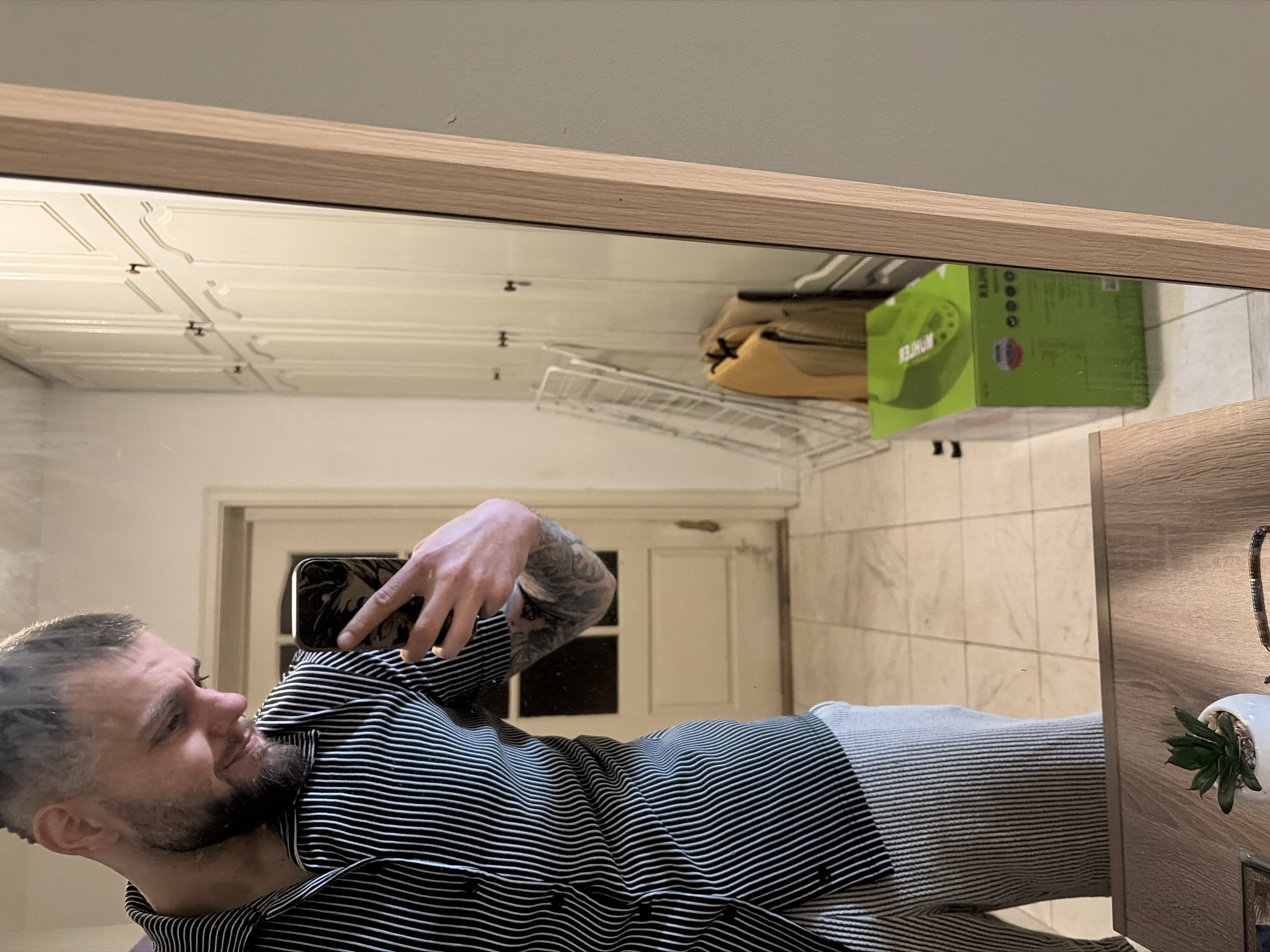 A man is taking a selfie in a mirror. The man has short, graying hair, a beard, and tattoos on his arm. He is wearing a striped shirt. The mirror is framed in a light wood and reflects a bathroom. The bathroom has white tile floors and a white door. There is a metal rack and a green box on a shelf above the door. There is a small potted plant on a table next to the mirror. The location cannot be determined from the image.