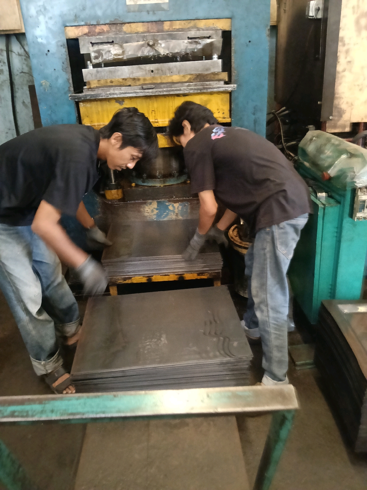 This is a medium shot, eye-level, indoor photograph of two young men working with sheet metal in what appears to be a factory or workshop in Belimbing, Indonesia. The men are both wearing dark t-shirts and denim jeans. The man on the left is leaning forward, his head down, as he reaches for a stack of flat metal sheets. He is wearing light-colored gloves. The man on the right is also wearing gloves and is reaching towards the same stack of metal sheets. A large, industrial metal press machine, painted blue and yellow, is visible in the background. The floor is concrete and appears somewhat wet or dusty. A stack of metal sheets is in the foreground, and other stacks of metal sheets are visible to the right, some covered in green plastic sheeting. The lighting is somewhat dim, suggesting an industrial setting.