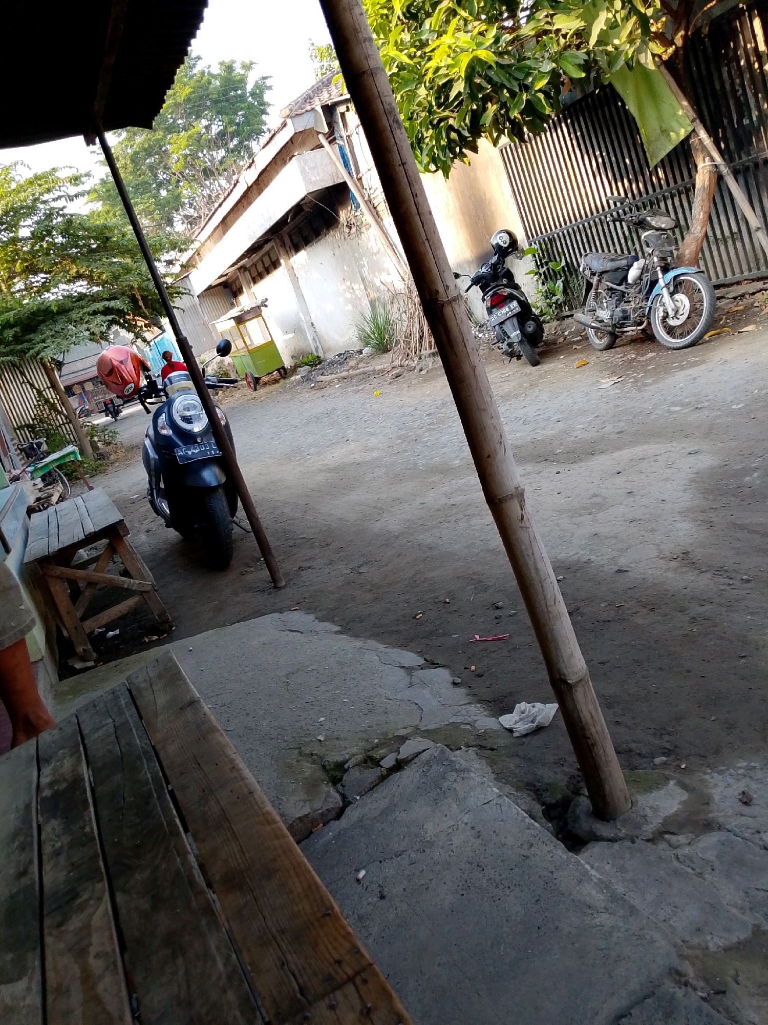 The image depicts a street scene in Gurah, Indonesia. In the foreground, a rustic wooden bench is partially visible on the left side, made of planks. A dirt road runs through the scene, with a concrete walkway on the right. A prominent bamboo pole stands vertically in the middle ground.
Several motorcycles are parked on the side of the road. Closest to the viewer, a dark-colored scooter is parked, with a red helmet on its seat. Further down the road, a black motorcycle is parked, and beyond that, a blue motorcycle. In the background, a simple building with a tiled roof and white walls is visible, along with some trees and a metal fence. A small green and yellow cart with wheels is also present. The overall impression is of a residential or commercial area with a relaxed, everyday atmosphere, likely during daylight hours given the lighting.