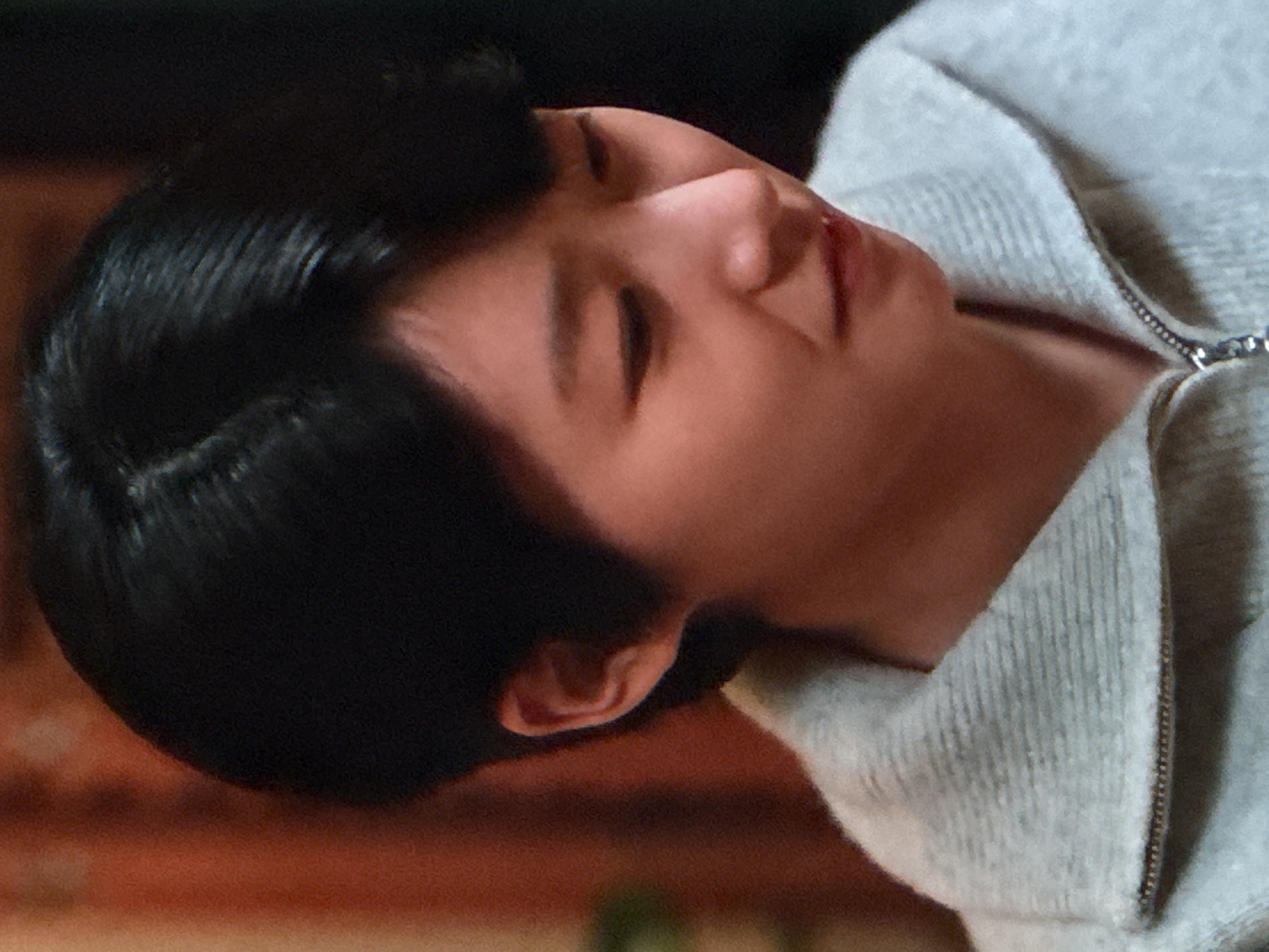 A close-up, head-and-shoulders shot shows a young Asian woman with short black hair and light skin. She is wearing a light gray, zippered sweater with a textured collar. Her head is tilted down and to her right, with her eyes looking downward and away from the camera. Her expression is one of sadness or distress, with her mouth slightly open and her lips slightly parted. The background is out of focus, with a reddish-orange textured surface on the left and a dark area on the right. The lighting appears to be dim, with a soft glow illuminating the woman's face from the front. The image is grainy, suggesting it may be a photograph taken in low light or a screenshot from a video. There are no discernible landmarks or environmental cues that would indicate a specific location in Unknown, Russia.