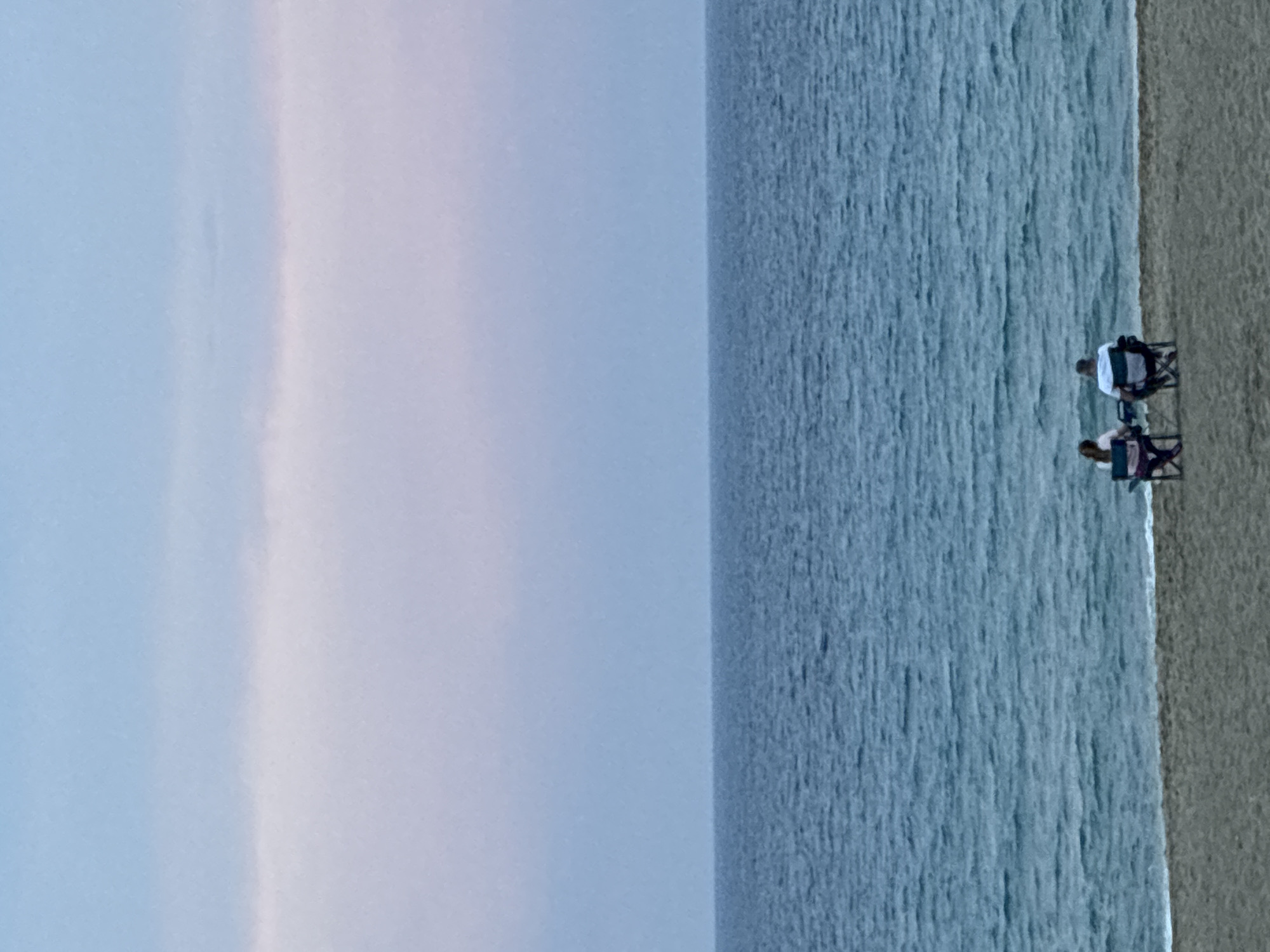 Two individuals, a male and a female, are seated in folding chairs on a sandy beach, facing a calm, blue ocean. The sky above is a gradient of pale blue and pink, suggesting it is either dawn or dusk. The beach is in Alanya, Turkey. The scene is tranquil and serene.