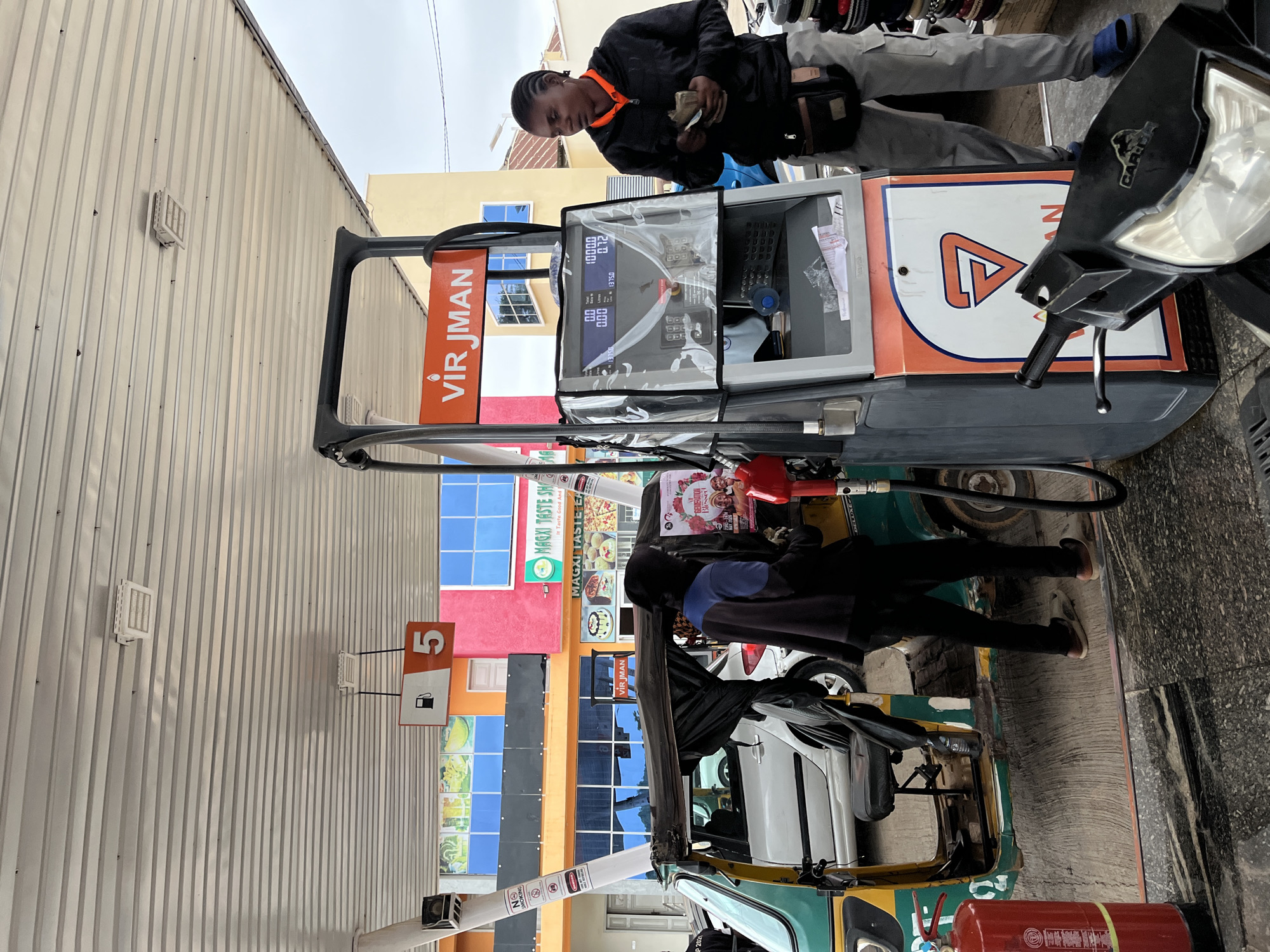 The image shows a scene at a petrol station in Jos, Nigeria. A man wearing a black jacket and grey pants is standing at a petrol pump, counting money. The pump displays the brand name "VIR JMAN" and indicates that it's pump number 5. A motorcycle is parked next to the pump. In the background, there's a building with colorful facades and a sign for "MAGXI TASTE SHAM". A three-wheeled vehicle, potentially a tricycle, is also visible near the pump with a fire extinguisher near it.