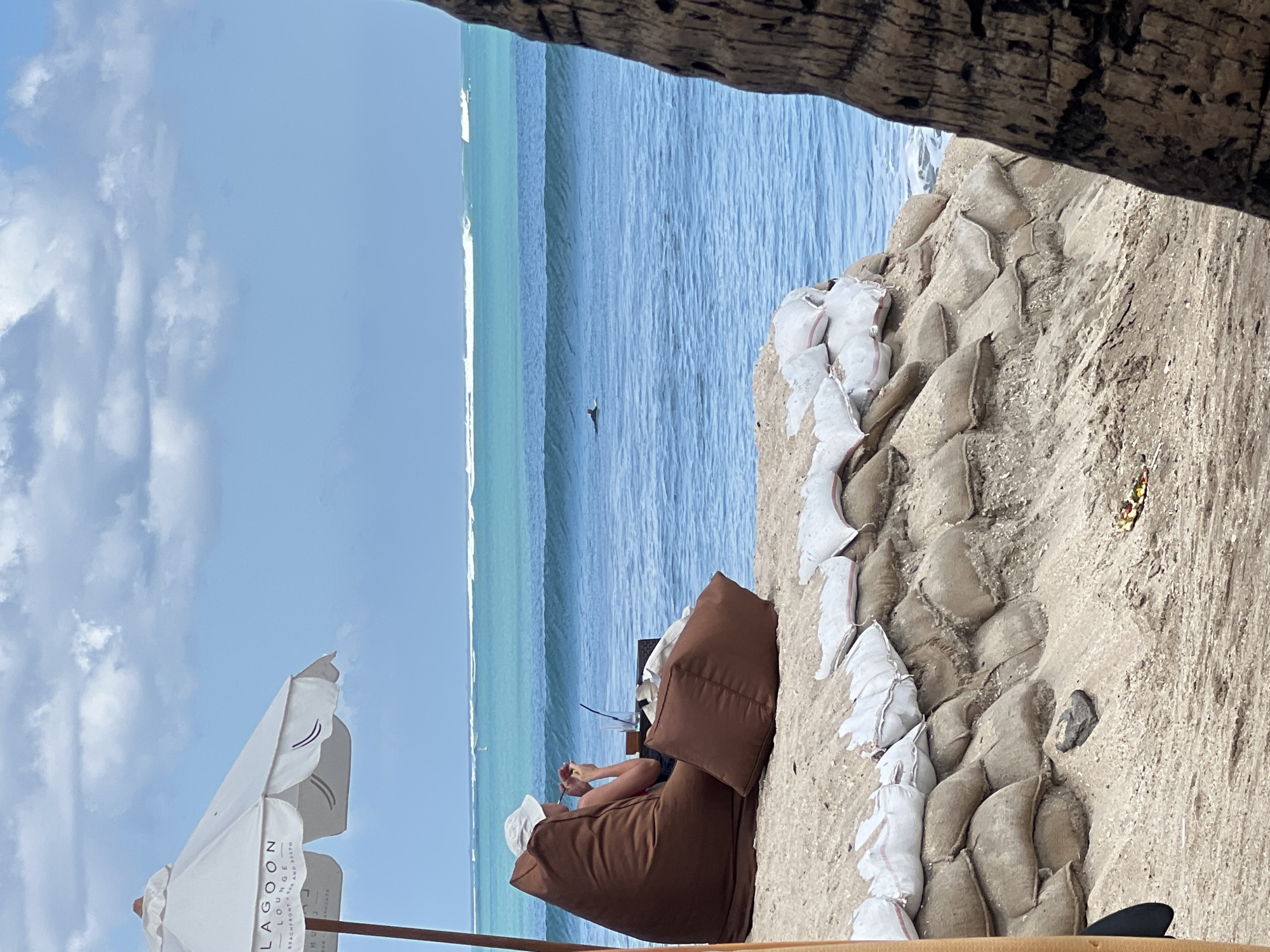 The image depicts a beach scene in Kuta, Indonesia. In the foreground, a person sits in a large brown beanbag chair on the sand, facing the ocean and holding a phone. A table with a drink sits beside them. Behind the person, sandbags are stacked in a tiered formation, acting as a barrier against the water. The ocean is visible with gentle waves, and a surfer can be seen in the distance. A white umbrella with the logo "LAGOON LOUNGE BEACHFRONT X BAR AND RESTO" is partially visible on the left side of the frame. The sky is partly cloudy. A portion of a palm tree trunk is visible on the far right, framing the scene.