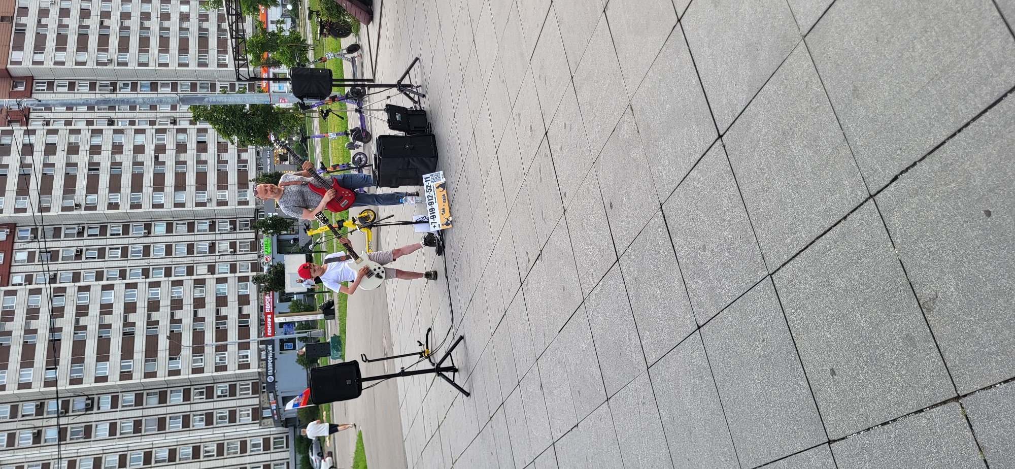 An outdoor, medium shot captures two males, one adult and one child, performing music on a city sidewalk in front of a large apartment building. The adult, on the right, is wearing a grey t-shirt and jeans, and playing a red electric guitar. The child, on the left, wears a white t-shirt and shorts, with a red baseball cap, and plays a white electric guitar. Both are standing, facing forward, with music equipment including speakers, amplifiers, and music stands around them. A sign with a phone number and text in Russian is placed on the ground near the child. In the background, several electric scooters are parked. The sidewalk is paved with large gray tiles, and the large building behind them is a light brown and beige multi-story structure with many windows. The sky is overcast.