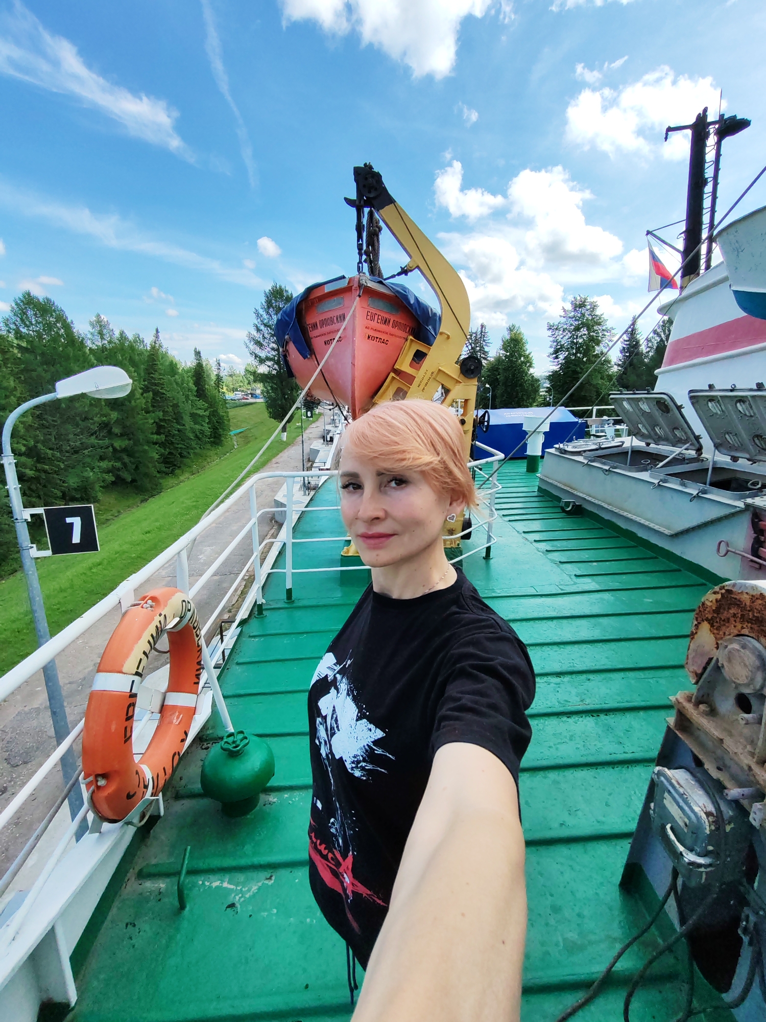 A woman with short, pinkish-blonde hair is taking a selfie on the deck of a boat. She is wearing a black t-shirt with a white and red graphic design. The deck is painted green. To her left, a life preserver is attached to a white railing, and a green buoy is visible. In the background, a red and orange life raft is suspended by a yellow crane. A mast with a Russian flag is also visible. Beyond the boat, there is a green, tree-covered embankment and a blue sky with white clouds. The location is identified as Belousovo, Russia.