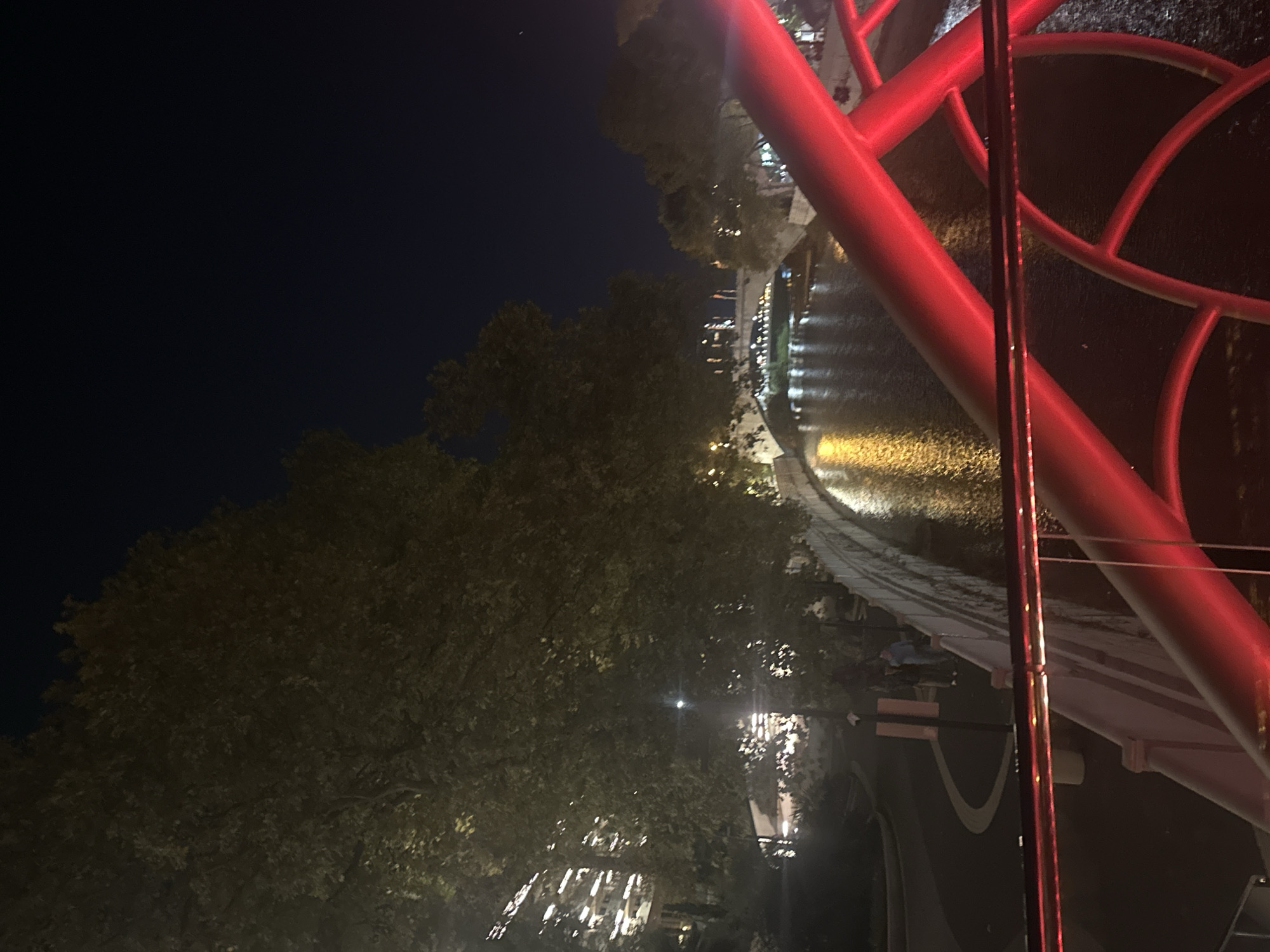 The image depicts a night view of the river promenade in Sochi, Russia, as seen from a bridge. A large tree with dense foliage dominates the left side of the frame, its leaves illuminated by ambient light, casting shadows on the background. To the left, a building with multiple floors and illuminated windows is visible, suggesting an urban setting. The river flows through the scene, its dark surface reflecting the lights from the city and bridges, creating a shimmering effect. A stone embankment lines the river, with some people standing on it, appearing to be engaged in conversation. A red-colored railing, part of the bridge the photo is taken from, is prominent in the foreground, with its vibrant color contrasting with the darker surroundings. Another bridge is visible in the distance, spanning the river, with lights illuminating its structure and the walkway above. The sky is dark, indicating it is nighttime.