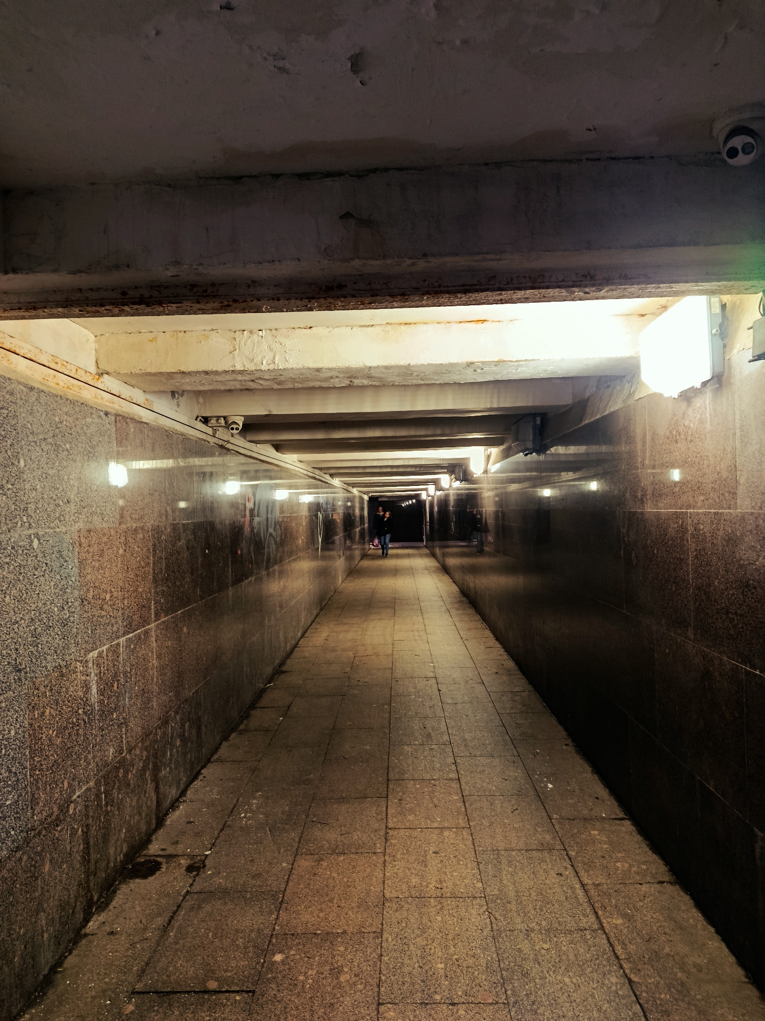 This is a ground-level, eye-level shot of a pedestrian underpass in Moscow, Russia. The underpass is illuminated by overhead fluorescent lights and has walls lined with dark, speckled granite tiles. The floor is paved with square concrete tiles. In the distance, two individuals are walking away from the camera. A security camera is visible mounted on the ceiling of the underpass, near the foreground. The overall impression is one of utilitarian infrastructure, common in urban environments.