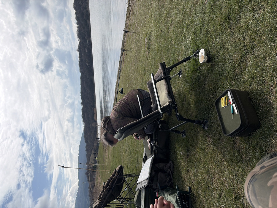 The image displays an outdoor fishing scene at a lakeside location near Cherna Gora, Bulgaria, under a partly cloudy sky. A male individual is seated in a dark green, adjustable fishing chair, leaning forward and engaging with items inside a large, open, dark green tackle bag positioned on the grass in front of him. He is wearing a camouflage-patterned hooded jacket and dark trousers. To his left, another large, dark green fishing chair or bedchair is partially visible. The bare forearm and hand of a second individual, holding a red object, are visible in the far left foreground.
On the grass in the immediate foreground, a small, rectangular dark green tackle box contains a silver knife, a blue tool, and a yellow container with small fishing tackle items. A silver mug is attached to the lower leg of the main subject's fishing chair. Along the water's edge to the right, at least three fishing rods are deployed from rod pods or banksticks. The body of water, likely a lake or reservoir, is calm with grey-blue tones, reflecting the sky. The far bank consists of a treed shoreline backed by rolling hills. Several indistinct figures are visible further along the distant bank, indicating other individuals engaged in fishing. The immediate ground is covered in short green grass.