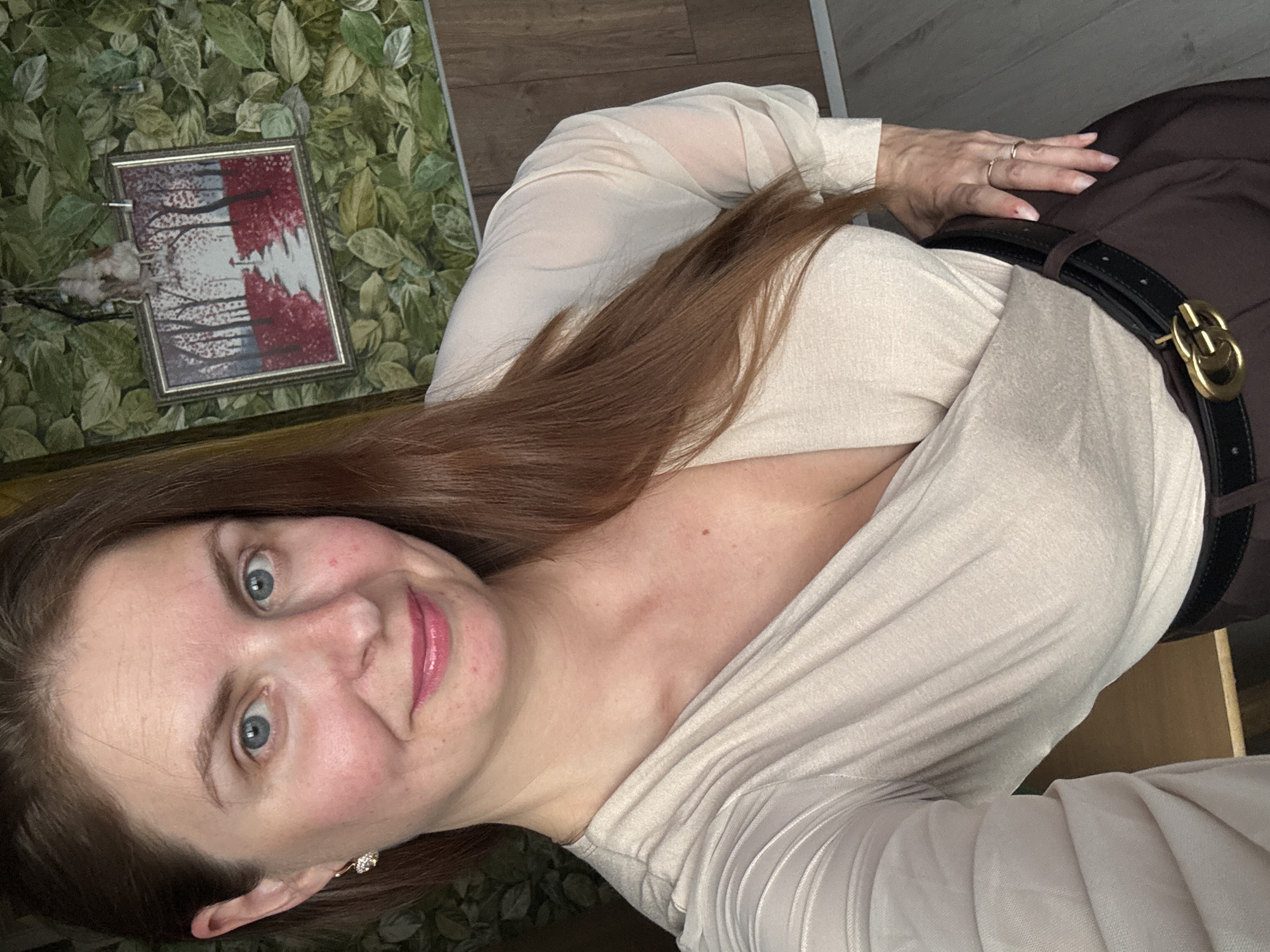 A woman with light skin, blue eyes, and long brown hair is posing for a selfie. She is wearing a light beige, low-cut wrap top and dark brown pants with a black belt. Her right hand is on her hip, and her left hand is resting on her thigh. The background features a wall covered in a green leafy pattern, a framed picture of a forest path, and wooden flooring. The image appears to be taken indoors, in what could be a residential setting. The specific location of Konakovo, Russia cannot be determined from the visual information provided in the image.