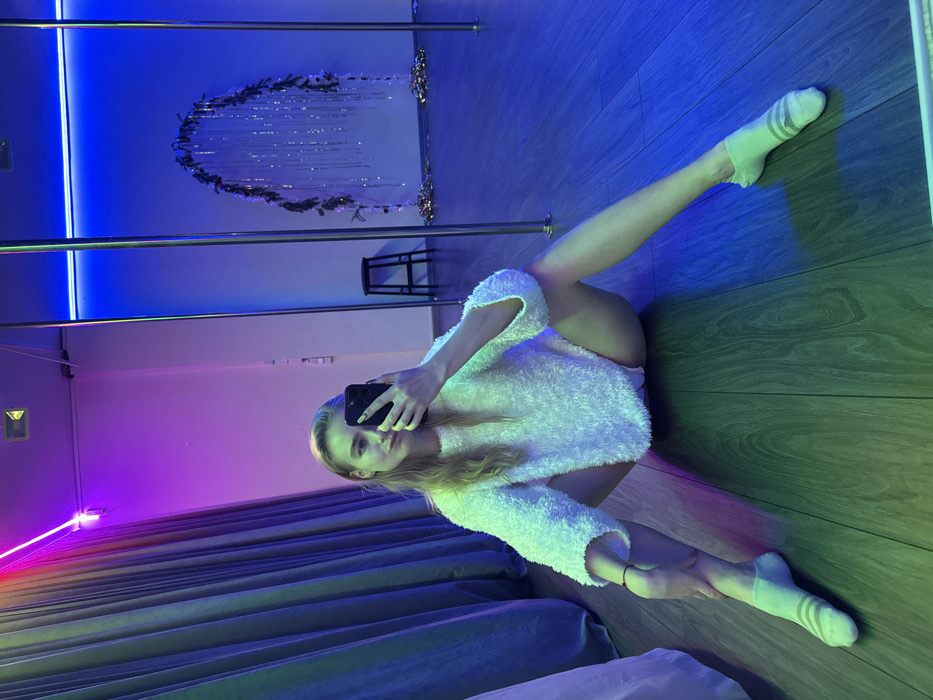 A woman in a white, fluffy sweater and socks is taking a selfie with a smartphone in a room with a reflective wall. The room has a neon blue light at the top and a purple light on the left side, giving it a vibrant atmosphere. A metal pole and a small stool are visible in the background, along with a curtain and what appears to be bedding. The floor has a wood grain texture and is illuminated with green light.