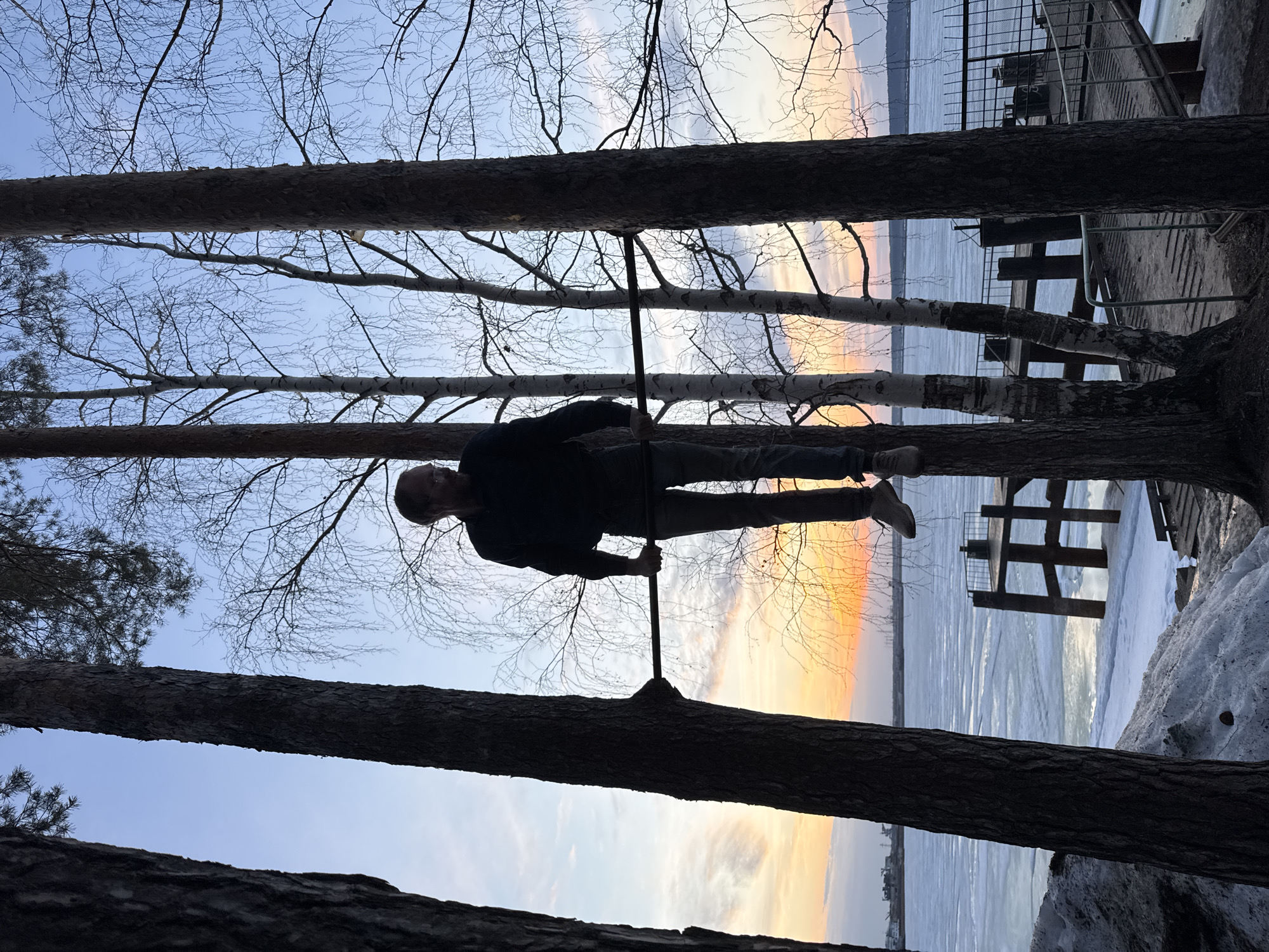 The image depicts a man performing a handstand on a pole, seemingly suspended between two trees. He is wearing a dark jacket and gray pants. The background features a view of the frozen Kama River in Chaikovsky, Russia. A partially frozen body of water is visible, along with a structure on the ice that appears to be a pier or some type of platform. Bare trees and a sunset-tinged sky are visible above. The surrounding environment includes snow on the ground.