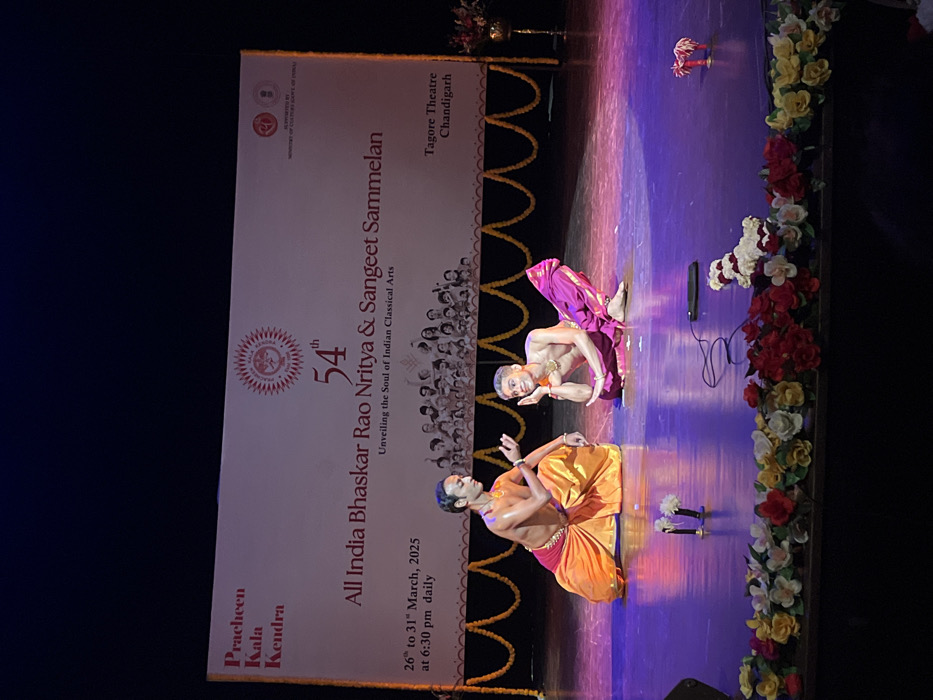 The image shows a performance at the Tagore Theatre in Chandigarh, India, located at the coordinates 30.733, 76.789. Two male dancers are performing on the stage. The dancer on the left wears an orange and gold costume and is in mid-gesture, while the dancer on the right wears a purple and gold costume and is sitting with his right hand on his chin. Both dancers are bare-chested and have dark hair. The stage is decorated with garlands of flowers and a black cable and cell phone lay in front of the dancers. A banner behind the dancers advertises "All India Bhaskar Rao Nritya & Sangeet Sammelan," the "54th Unveiling the Soul of Indian Classical Arts," and "26th to 31st March, 2025 at 6:30 pm daily." The banner also lists the venue as "Tagore Theatre Chandigarh." The backdrop behind the banner is a dark, blurred curtain. The lighting is dim, casting a bluish hue over the stage.