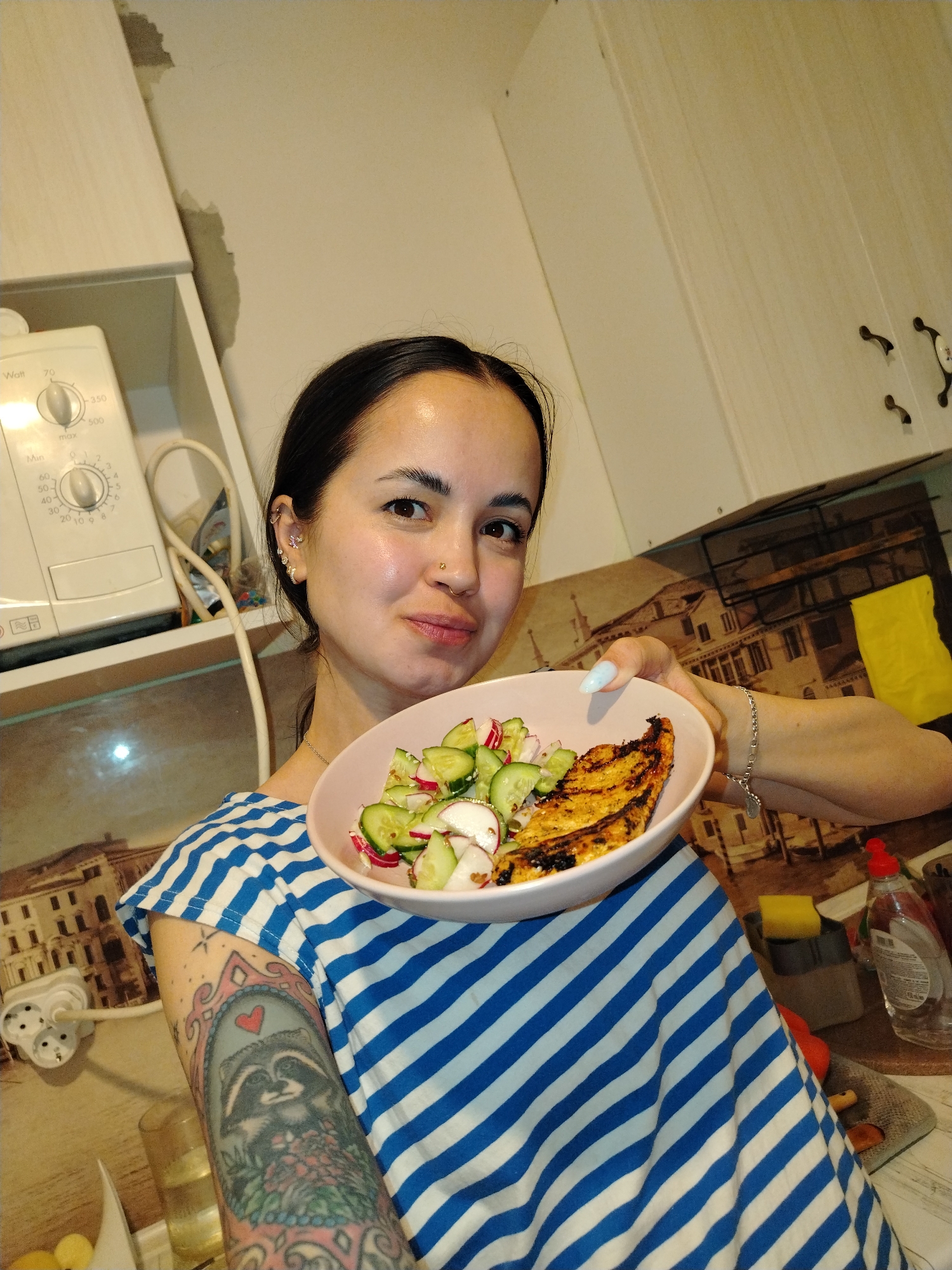 A young woman is depicted in a chest-up shot within a kitchen setting, likely in Noviy, Russia. She has dark hair pulled back and wears a blue and white striped top. Her expression is a soft, pleasant smile, and she features multiple ear piercings with intricate jewelry and a nose piercing. She holds up a light pink bowl containing a meal of grilled protein (possibly chicken or fish) and a fresh salad composed of sliced cucumbers and radishes.
Her visible left arm is adorned with a colorful sleeve tattoo, prominently featuring a raccoon within an ornate frame, topped by a red heart. The background reveals typical kitchen elements, including white upper cabinets with dark handles. To her left, a white kitchen appliance, possibly a food processor or blender base, is visible on a shelf. Its dials display text such as "Watt" with settings "70, 350, 500 max" and "Min" with time markings "60, 50, 40, 30, 20, 10" and another dial with numbers "9, 8, 7, 6, 3, 2, 1." A power cord snakes from the appliance, and below it, a multi-socket adapter is plugged into a wall outlet. The kitchen backsplash features a printed mural of an old European cityscape, possibly Venice, complete with buildings and water. On the counter to her right, a yellow sponge and a bottle of dish soap are partially visible. The scene is illuminated by indoor lighting.