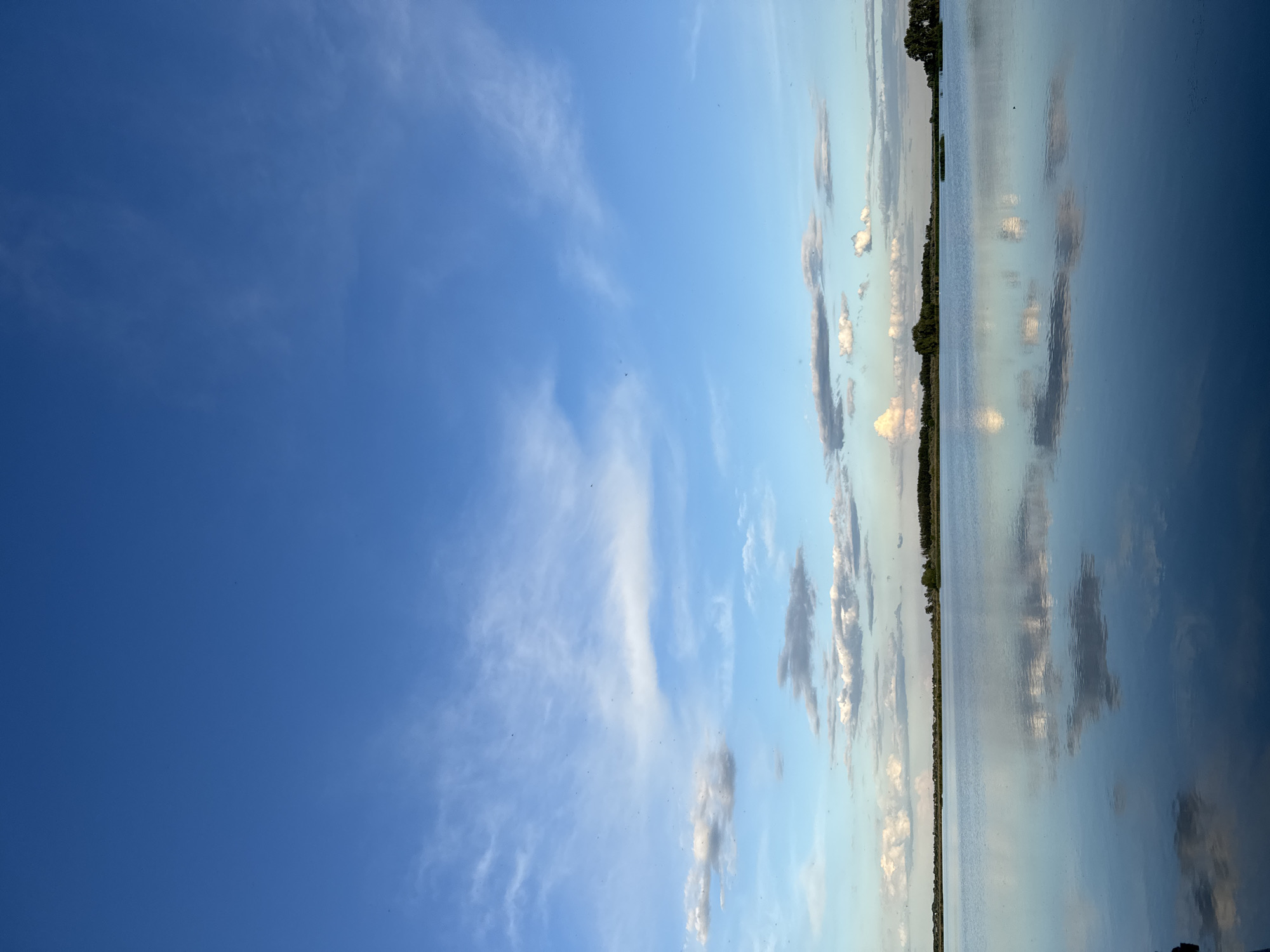 The image shows a landscape with a body of water, possibly a lake or river, occupying the right side of the frame. The water reflects the sky and clouds above, creating a mirror-like effect. A thin strip of land with trees runs along the horizon. The left side of the image is dominated by a blue sky with wispy clouds. The location is Unknown, Unknown.