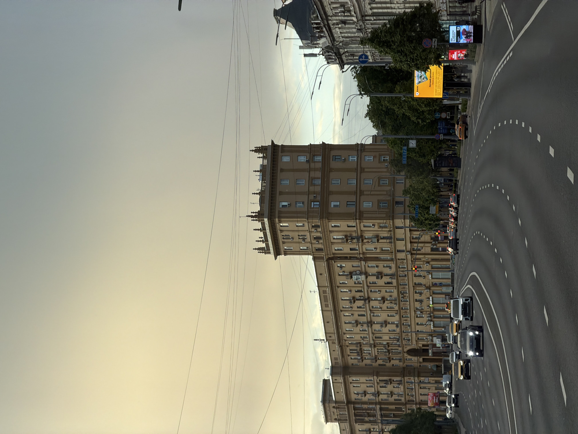 The image captures a street scene in Moscow, Russia. The prominent architectural feature is a tall, multi-story building with a classical design, beige facade, and repetitive window patterns. A road runs adjacent to the building, curving slightly into the distance. Vehicles, including cars and SUVs, occupy the road. Streetlights and advertising billboards line the sidewalk. The sky is overcast, with a yellow-grey hue. Utility wires are visible overhead. Trees are interspersed along the sidewalk and in the background.