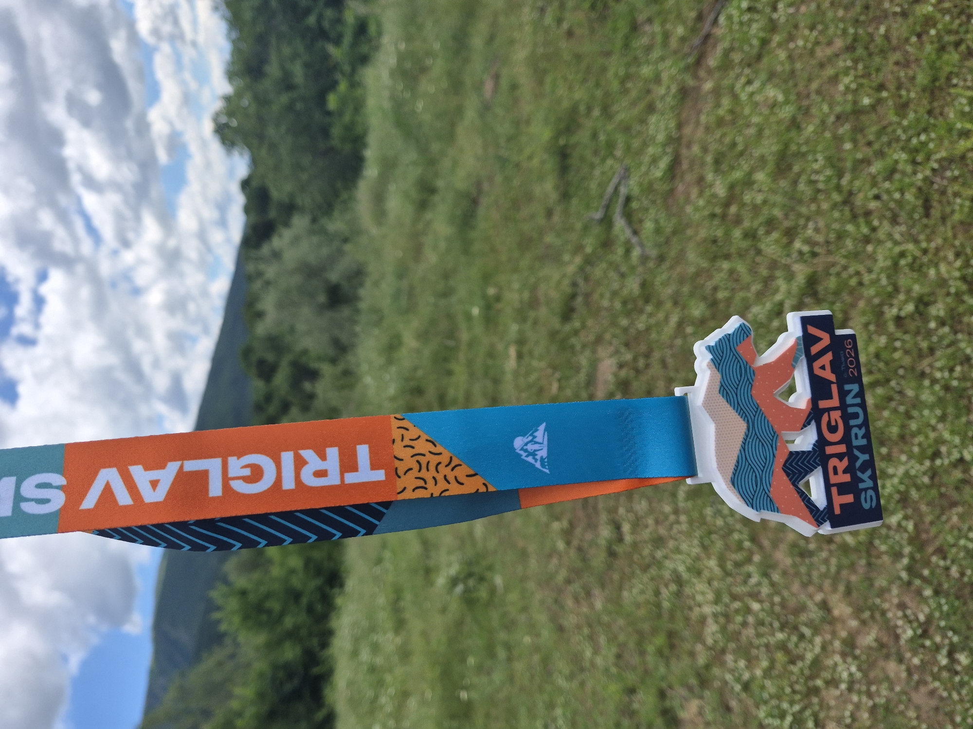 A close-up, shallow depth-of-field shot shows a medal for the "Triglav Skyrun 2026" event hanging from its ribbon. The ribbon is orange and turquoise with a black chevron pattern and abstract orange and black shapes. The medal itself is shaped like a bear with colorful mountain-like patterns and the event name and year prominently displayed. The background is a blur of green grass with scattered white flowers, and distant green trees and hills under a cloudy blue sky. The location is identified as the city of Tazha, Bulgaria. There are no people or animals visible in the image.