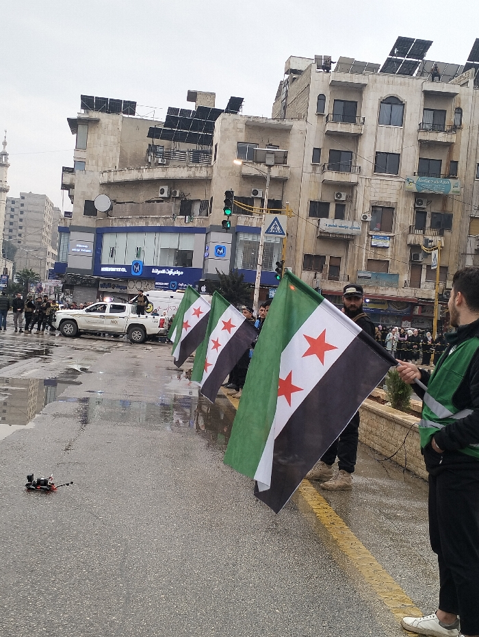 A street scene shows individuals holding Syrian flags. A drone sits on the wet pavement near the flag bearers. A white vehicle with writing on its side is parked nearby, surrounded by other people. Buildings line the background, many adorned with solar panels. A tall minaret is visible in the distance, adding to the urban setting.