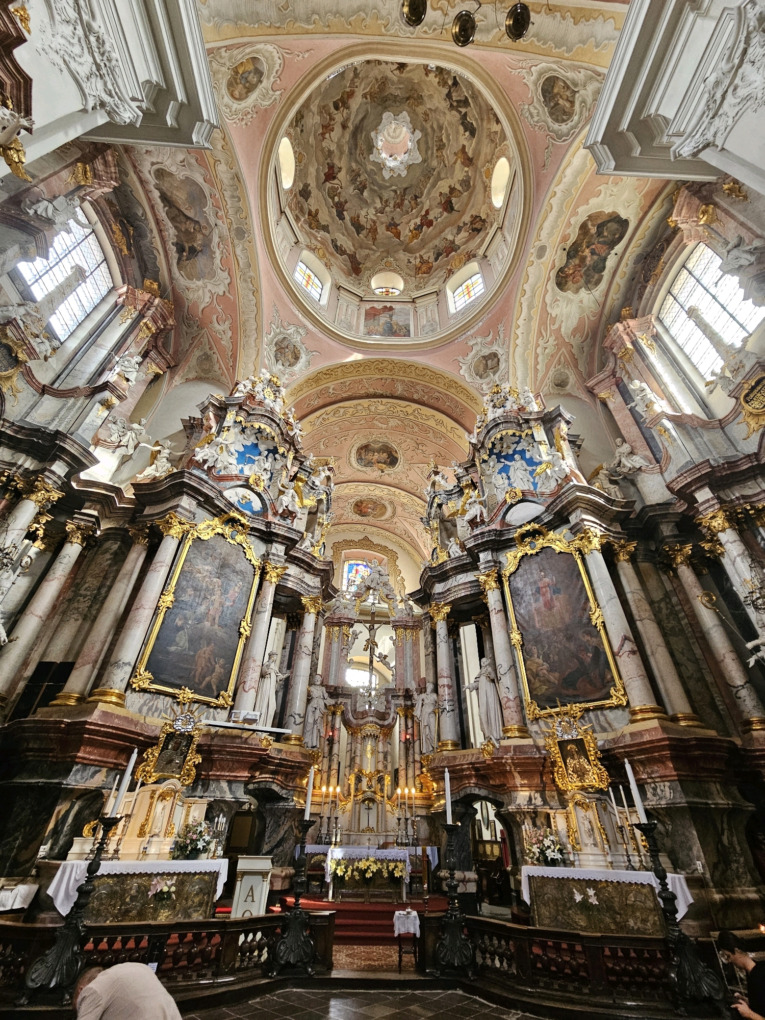 The image shows the interior of St. John's Church (Šv. Jonų bažnyčia) in Vilnius, Lithuania, looking towards the altar. The church features elaborate Baroque architecture with ornate columns, gilded decorations, and religious paintings on the walls and dome. The dome is particularly striking, adorned with frescoes depicting religious scenes and figures. The altar area is richly decorated with sculptures, paintings, and floral arrangements. Several individuals are visible in the lower part of the frame, including one person kneeling in the foreground. The lighting suggests it is daytime, with natural light entering through the windows and artificial lighting from the church's fixtures.