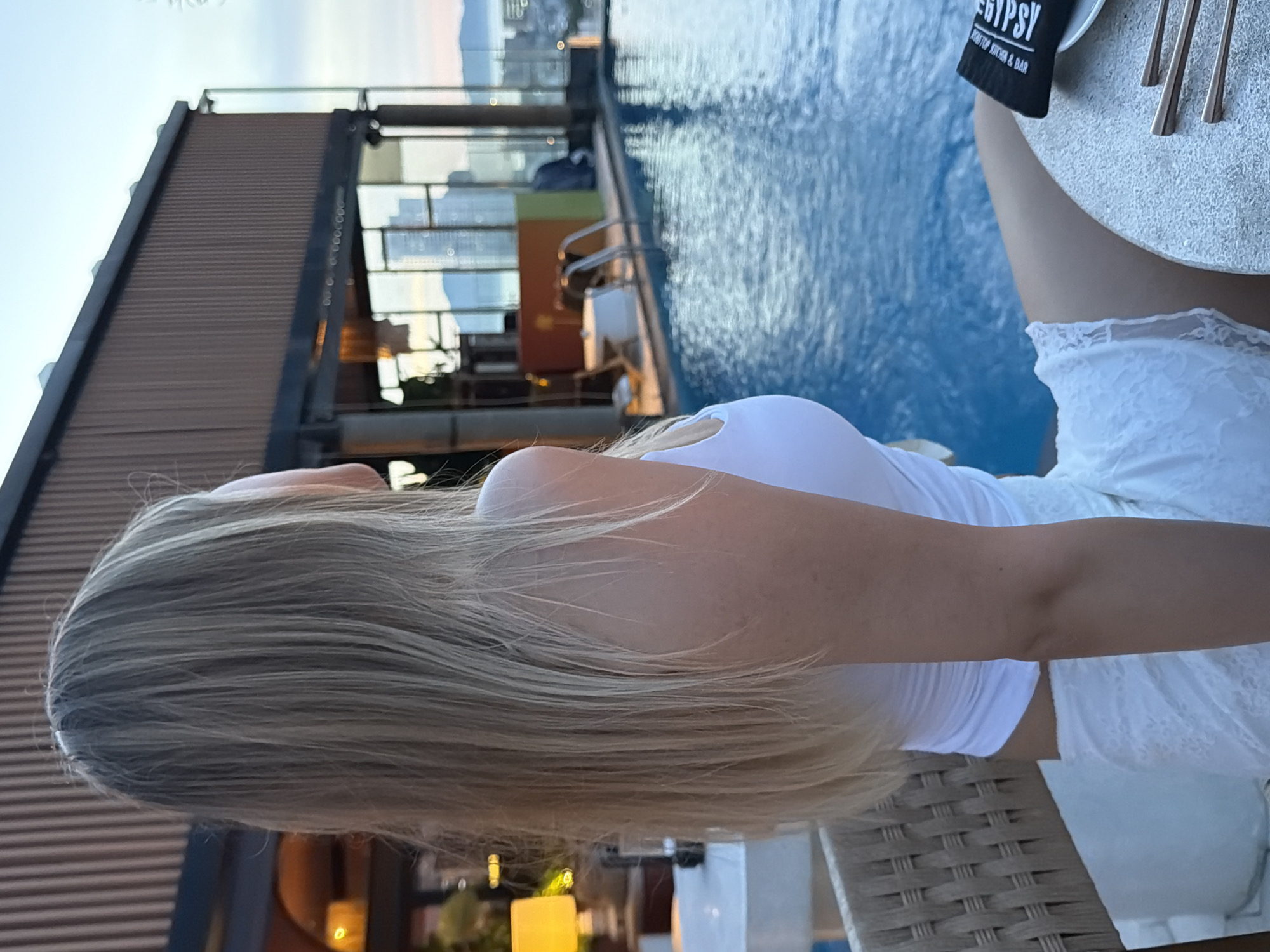 An outdoor rooftop pool area in Đà Nẵng, Vietnam, is featured in this image. A woman with long blonde hair is seated, facing away from the camera, looking out towards the pool and city skyline. She is wearing a white tank top and a white lace skirt. The pool is a vibrant blue, with a ladder leading into it. In the background, a modern building with large glass windows overlooks the city, with mountains visible in the distance. The sky is a soft, overcast blue. A table with cutlery and a black napkin with "Gypsy" printed on it is visible in the foreground.