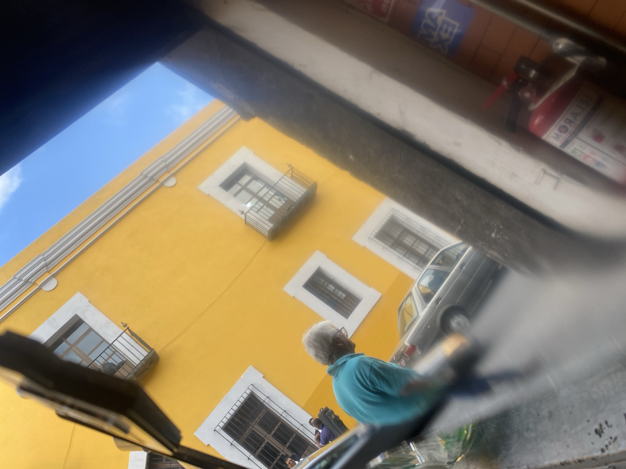 The image, taken from a low angle, captures a scene in Puebla, Mexico. A vibrant yellow building with white-framed windows and wrought-iron balconies dominates the background. A senior man with gray hair wearing a blue shirt stands in the foreground. A silver sedan is adjacent to him. Above, a blurry fire extinguisher is mounted on the wall. The overall setting is a bustling street scene, but the focal point is the architectural detail of the yellow building and the people.