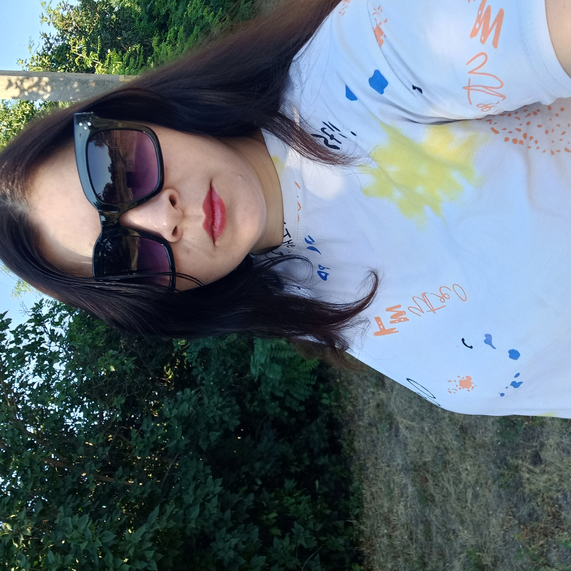 A medium shot shows a woman wearing large, black, square-framed sunglasses with purple lenses, dark lipstick, and a white t-shirt with colorful abstract paint splatters and some handwritten text. She has long, dark hair. She is standing outdoors, with green foliage and trees in the background, and dry grass and a utility pole to her right. The image appears to be taken in the city of Klyntsi, Ukraine, as indicated by the provided context.