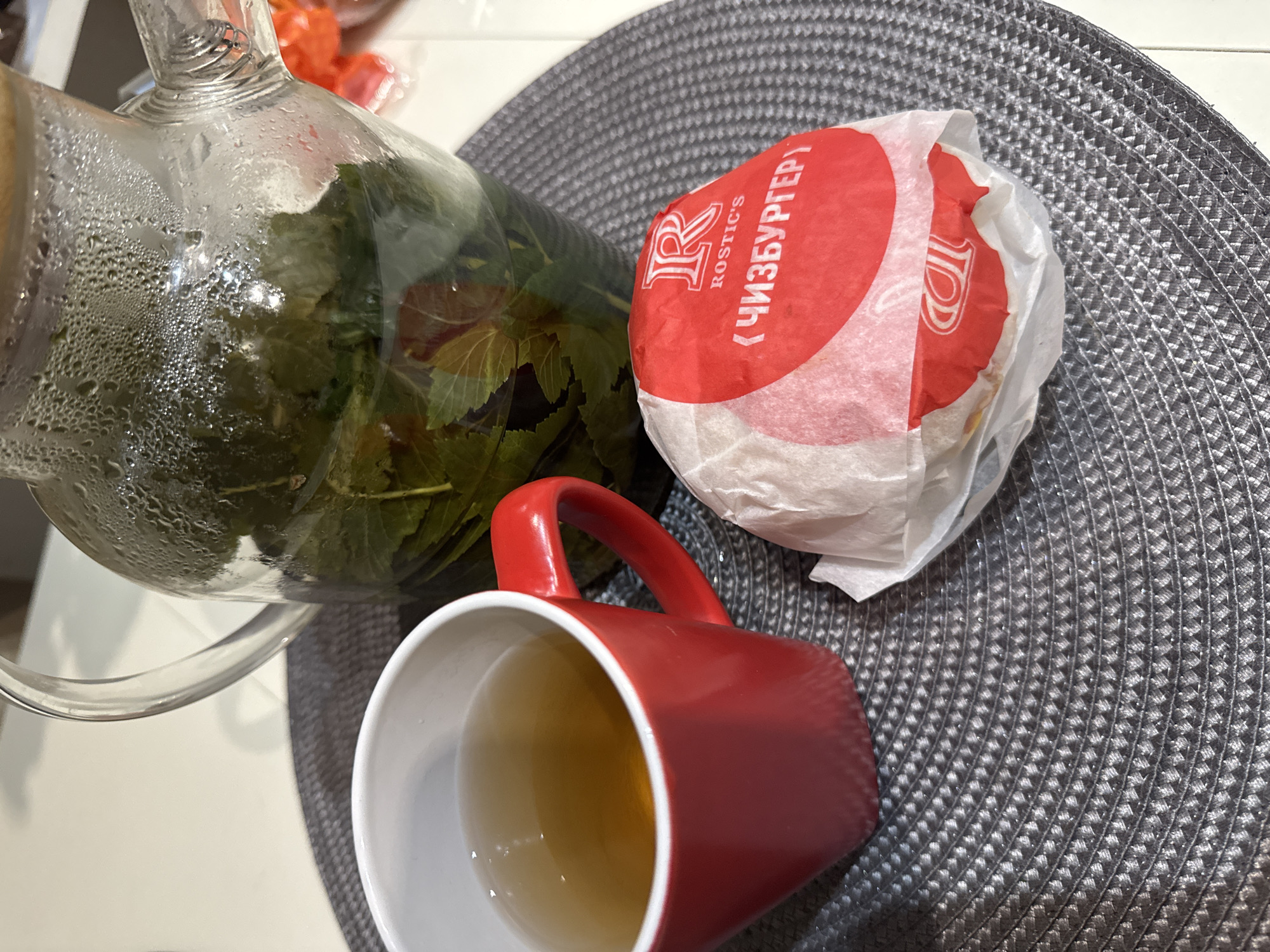 In the city of Chaikovsky, Russia, a close-up, eye-level shot captures a still life arrangement on a circular gray placemat. To the left, a vibrant red mug is filled with a light brown liquid, likely tea. Beside it, a glass pitcher, slightly fogged with condensation, contains a brewed herbal tea with visible green leaves. In the foreground, a single burger, wrapped in white paper with a prominent red circular sticker featuring a white "R" logo and Russian text, rests on the placemat. The background is blurred, showing a light-colored surface and hints of orange objects, possibly packaging, to the right. The scene suggests a moment of simple refreshment, with the tea and burger indicating a meal or snack.