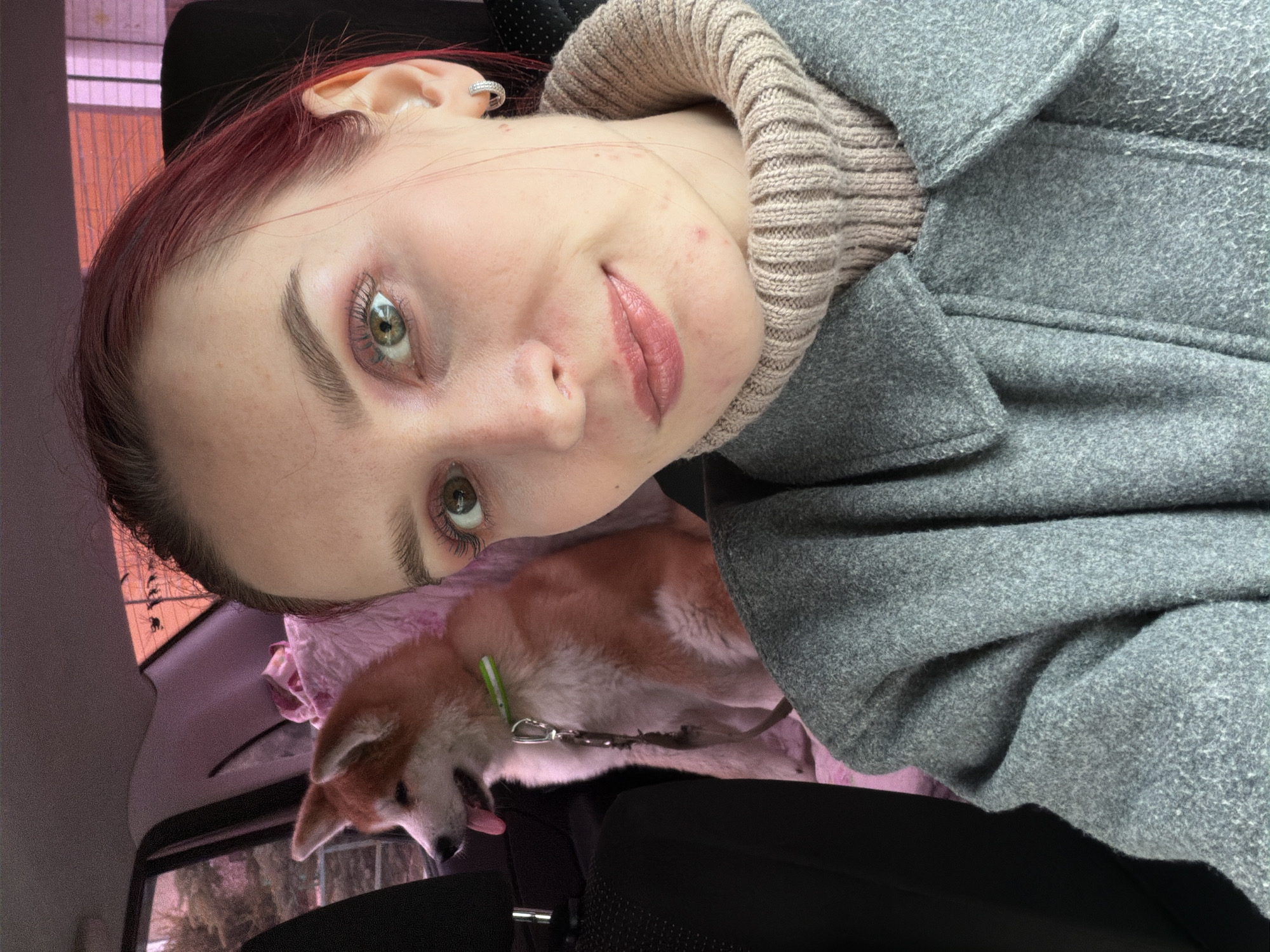 This media file is a close-up selfie-style photograph, rotated approximately 90 degrees counter-clockwise, featuring a person and a small dog, taken likely inside a car in Chaikovsky, Russia.
The person, who dominates the upper left portion of the frame (due to rotation), has fair skin, reddish-brown hair, and green eyes. They are wearing a light beige ribbed turtleneck sweater beneath a grey, wool-textured coat. Subtle makeup highlights their eyes with mascara and their lips with a rosy tint. A silver-colored hoop earring is visible in their ear. Their expression is a gentle, pleasant smile, and a few blemishes are visible on their skin.
Below and to the right of the person is a small dog, possibly an Akita or Shiba Inu mix, with fawn and white fur. It wears a light green collar with a leash attached and is resting on a soft pink blanket or fabric, with its tongue slightly extended.
The background elements suggest an indoor setting, specifically a car interior, with visible headrests and seat fabric. Through the car windows, hints of an outdoor scene can be discerned, characterized by subdued, possibly overcast, lighting. The grey coat worn by the person suggests cooler weather conditions. No visible text is present in the image. The overall mood is calm and personal.