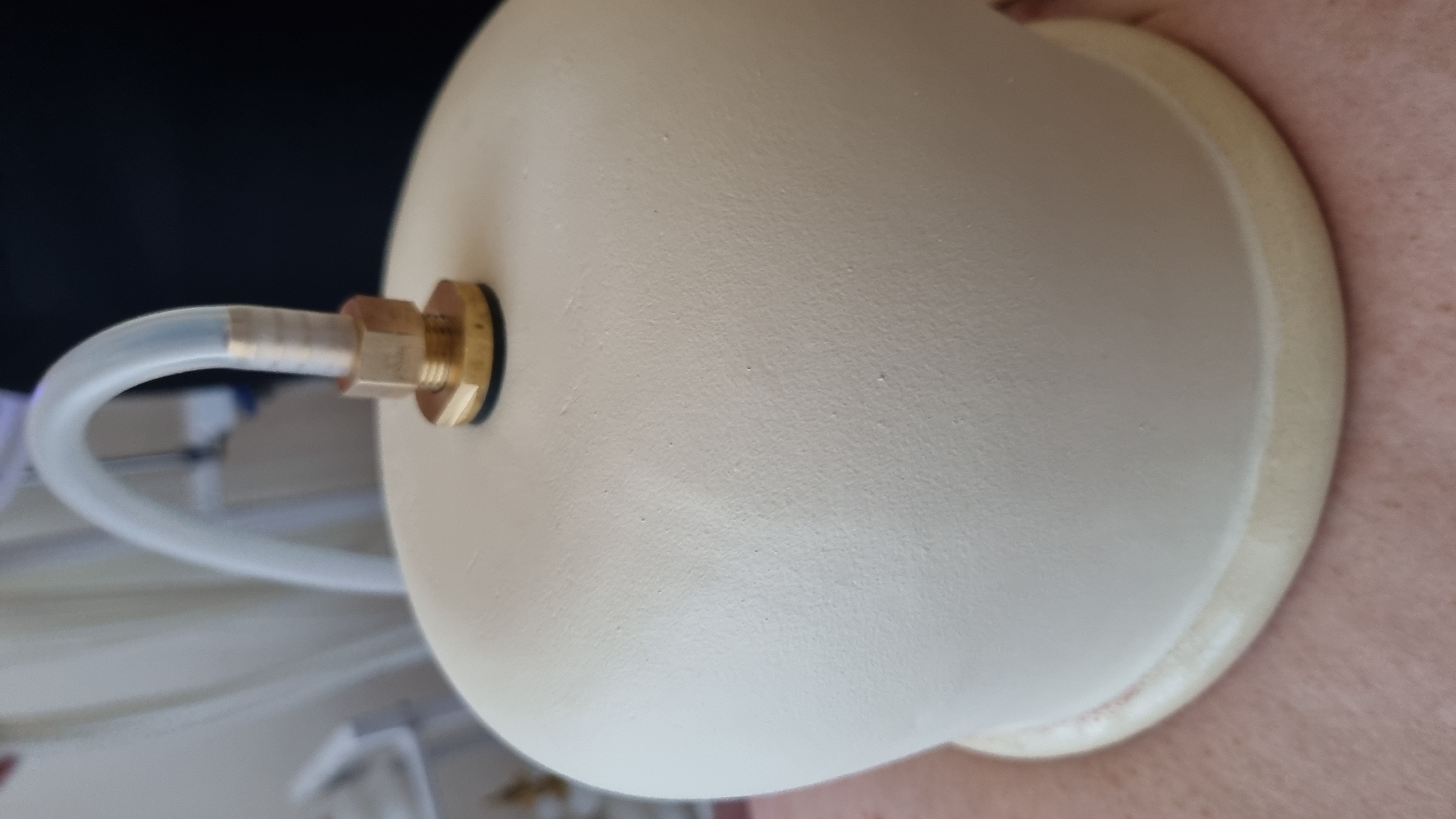 The image shows a close-up of a medical device, likely a vacuum cup, being applied to a person's skin. The device is a large, cream-colored, dome-shaped object with a ribbed, light yellow edge at its base. Attached to the top of the dome is a brass fitting, through which a clear, flexible tube extends upwards. The tube appears to be connected to medical equipment, as suggested by a blurred background with what looks like medical machinery. The skin visible beneath the device shows a light pink tone with subtle texture. The scene is brightly lit, and the focus is sharp on the device and its immediate surroundings. The image does not provide any information about the city of Mukachevo, Ukraine.