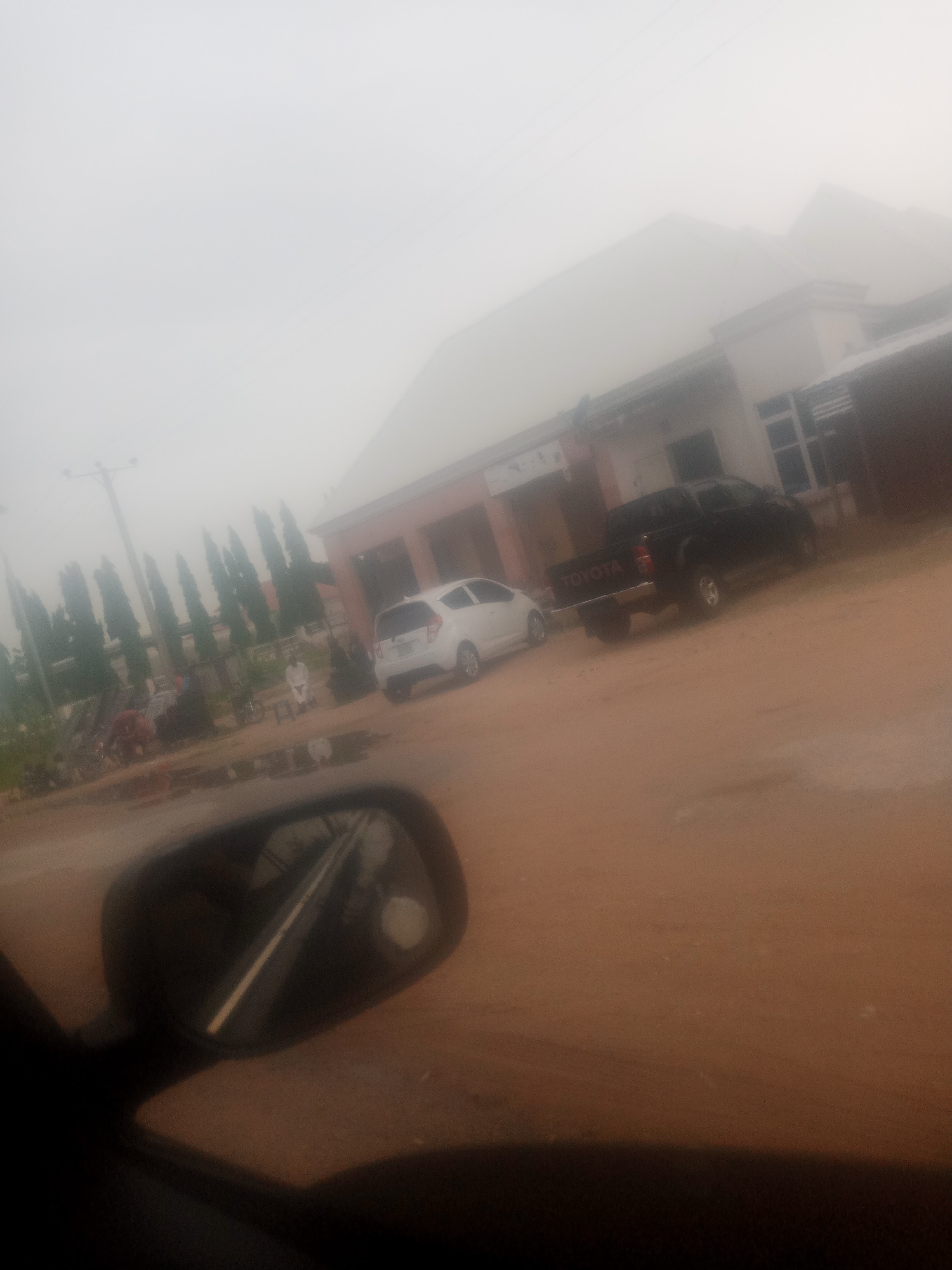 The image depicts a street scene in Jalingo, Nigeria, characterized by overcast weather, potentially fog or mist, which obscures the sky and softens the overall lighting. In the foreground, a portion of a car's side mirror is visible, reflecting a road with a white line down the center, suggesting the perspective of a driver.
The middle ground of the image features a dirt or unpaved road with several vehicles. To the left, a white hatchback is parked. Further back, a black Toyota pickup truck is parked on the right side of the road. Behind these vehicles, a light-colored building with a prominent roofline is situated. The building appears to have multiple windows.
To the left of the building, a row of tall, slender trees stands, their forms somewhat indistinct due to the atmospheric conditions. There are also signs of human activity; a person in a white garment is seated on a bench, and other individuals and possibly market stalls are visible in the background, though details are limited by the low visibility. The ground is primarily composed of brown dirt, with some patches that appear damp or reflective, possibly puddles. The overall impression is of a somewhat hazy and muted urban or semi-urban environment.