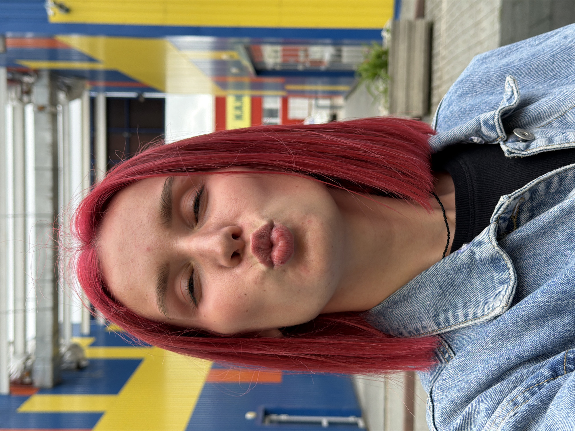 A young woman with bright red, shoulder-length hair is depicted in a close-up selfie, making a pouting or kissing gesture with her lips pursed forward and eyes squinted. She wears a light blue denim jacket over a black ribbed top and a thin black beaded necklace. The background is an outdoor urban or commercial area in Chaikovsky, Russia, featuring buildings with prominent blue, yellow, and red/orange rectangular panels. Partial Cyrillic lettering, specifically "К.А.М." (K.A.M.), is visible on a yellow section of a distant building to the right. The ground is paved, and planters containing greenery are present. The sky is overcast, providing diffuse natural lighting.