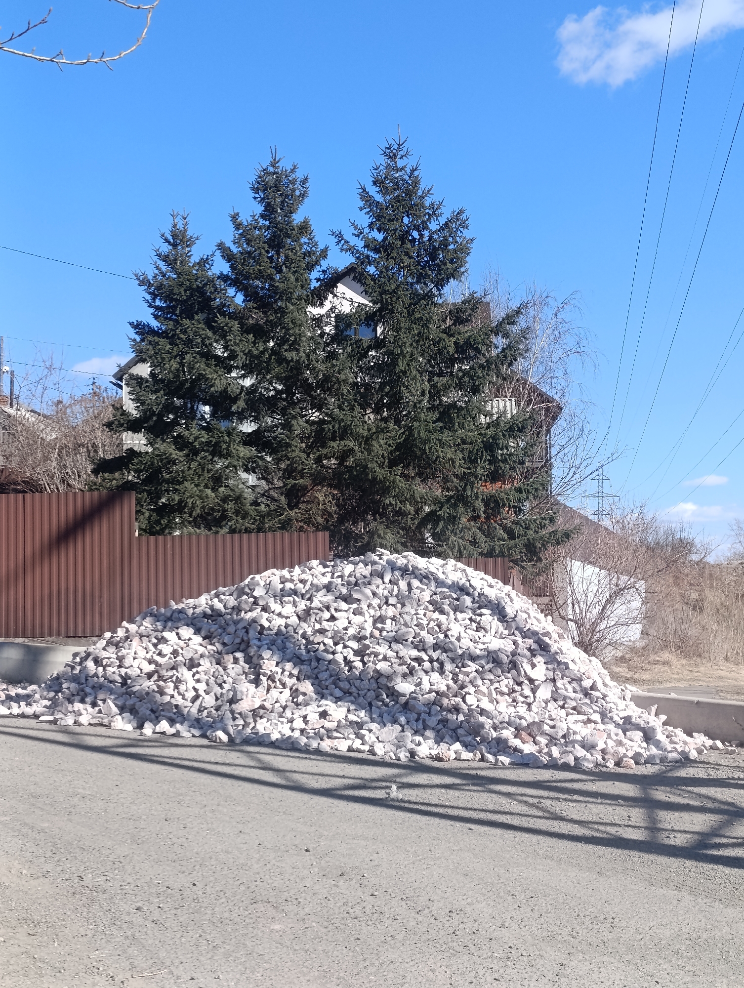 The image depicts a large pile of white rocks or gravel on a paved road in what appears to be a residential area. In the background, there is a house partially obscured by three evergreen trees, and a brown corrugated metal fence to the left. Power lines are visible against a clear blue sky. The photograph was taken in Krasnoyarsk, Russia.
