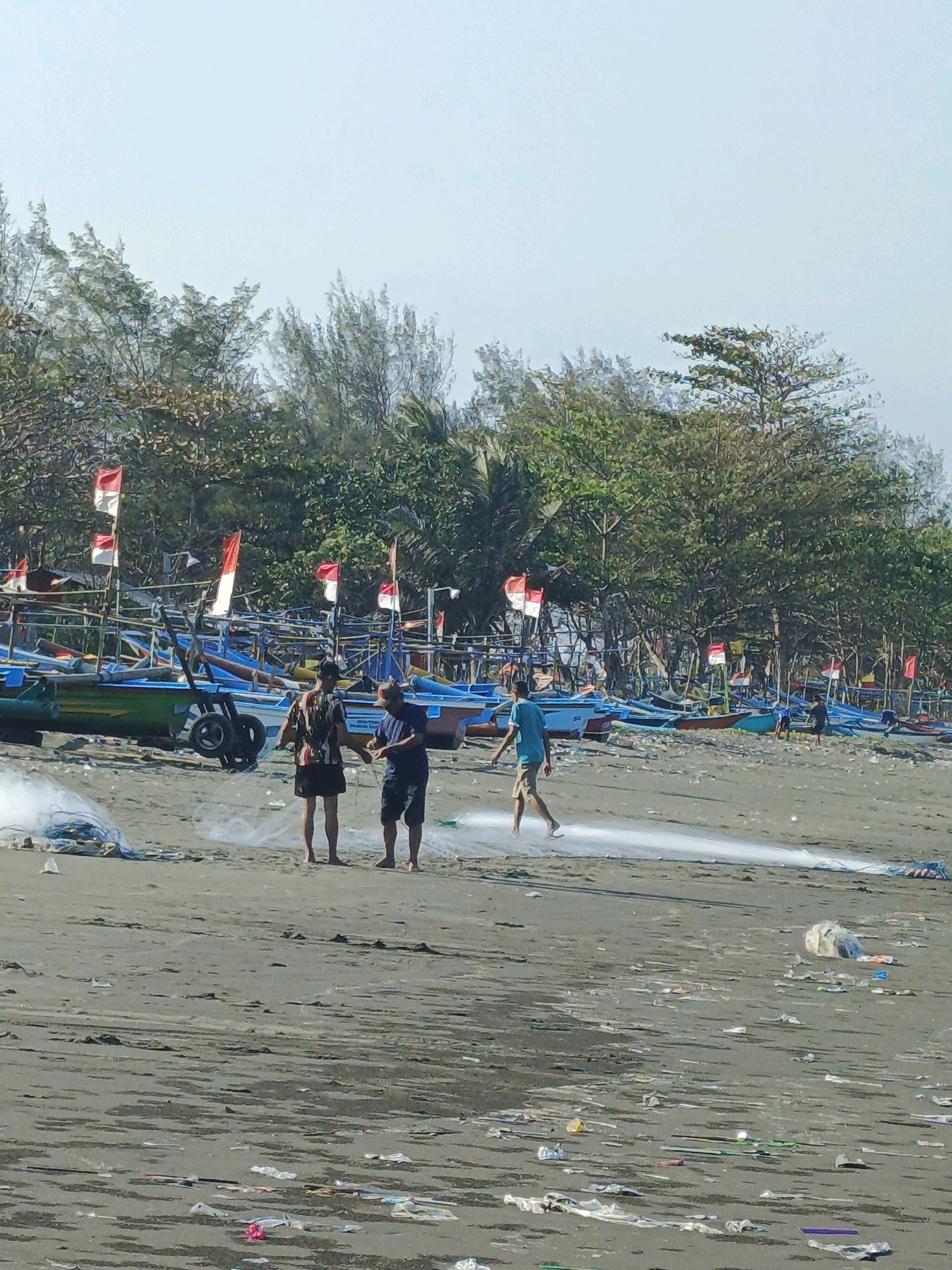 The image depicts a beach scene in Kelurahan Cilacap, Kecamatan Cilacap, Indonesia. In the foreground, a sandy beach is littered with various forms of trash, including plastic bags and bottles. Three individuals are present on the beach. Two men are standing together, appearing to be working with a fishing net that is spread out on the sand. A third person, possibly a child, is walking in the background, also near the fishing net. Behind the people and the net, a line of colorful fishing boats are pulled up onto the shore. Red and white Indonesian flags are prominently displayed on poles attached to the boats, suggesting a national celebration or event. A dense line of trees forms the background of the scene, with a clear, bright sky above.