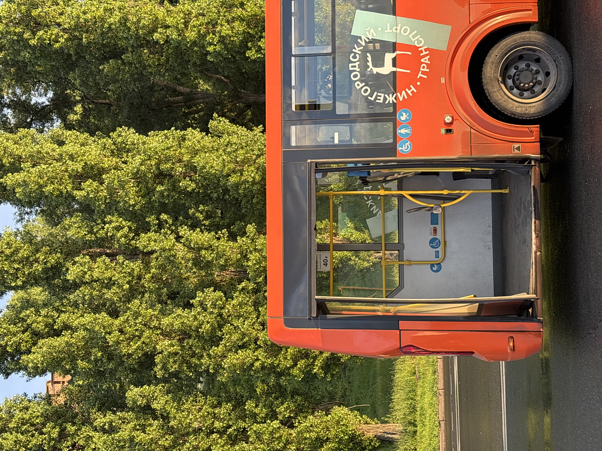 The image shows the side of a red bus parked next to a green tree-filled area. The bus is a public transport vehicle, indicated by the text "НИЖЕГОРОДСКИЙ ТРАНСПОРТ" (Nizhny Novgorod Transport) and a graphic of a deer on its side. There are also universal symbols for accessibility, such as a wheelchair user icon and a person icon, suggesting the bus is equipped for passengers with disabilities. The bus has large windows and a visible entrance area with yellow handrails. The time of day appears to be daytime, with bright sunlight illuminating the scene, creating a pleasant and warm atmosphere. The setting seems to be a street or a parking area, likely in an urban or suburban environment, given the presence of the bus and the paved road. The lush green trees suggest it's a season with abundant foliage, possibly spring or summer. There is also a sign inside the bus with "40 р" written on it, which likely indicates a fare or route number. The overall impression is that of a functional public service vehicle in a natural, sunlit environment.