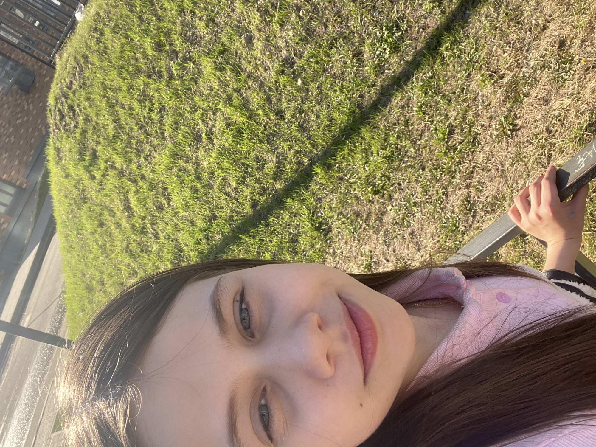 A young woman is depicted taking a selfie outdoors in Izhevsk, Russia. The image is rotated 90 degrees counter-clockwise. Her face, seen from a slightly low angle, features light skin, light blue-green eyes, and a gentle, natural smile. She has long, dark brown hair that falls around her face. She is wearing a light pink collared shirt, possibly with a subtle woven pattern, and a portion of a striped dark and light garment, likely a cardigan or sweater, is visible on her left shoulder.
Her right hand is seen in the lower right quadrant, holding onto a dark, possibly metal, railing or fence. There are some indistinct white markings or graffiti on the railing. The background reveals a sunny outdoor scene. The ground is covered with a mix of vibrant green grass and patches of drier, light brown grass. Long, distinct shadows are cast across the grass, indicating direct sunlight and suggesting it is either morning or late afternoon. To the left of the frame, a paved area or road runs along the edge. In the upper left background, part of a multi-story brick building with several windows and an exterior unit, possibly an air conditioner, can be seen. The weather appears clear and sunny.