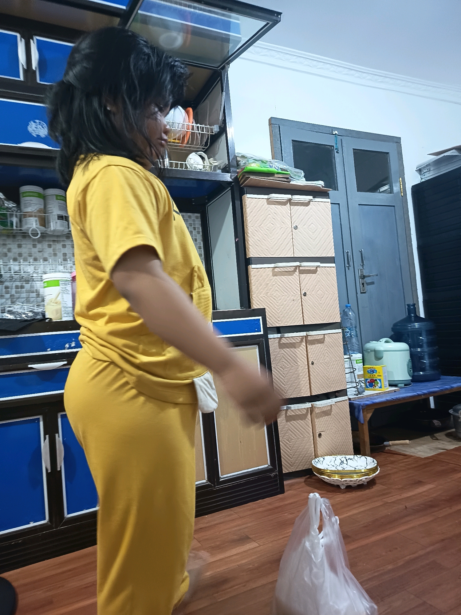 The image depicts an indoor scene, likely a kitchen or dining area, with a woman in the foreground wearing a yellow t-shirt and yellow pants. She is facing away from the camera, with her right arm extended and blurred due to motion. Behind her, there are kitchen cabinets, shelves with containers, a blue door, and various household items including water dispensers, a rice cooker, and a stack of plates. The floor is made of polished wood. The overall impression is of a domestic setting. The image does not provide any specific clues that would allow for identification of its location within Banjarmasin, Indonesia.