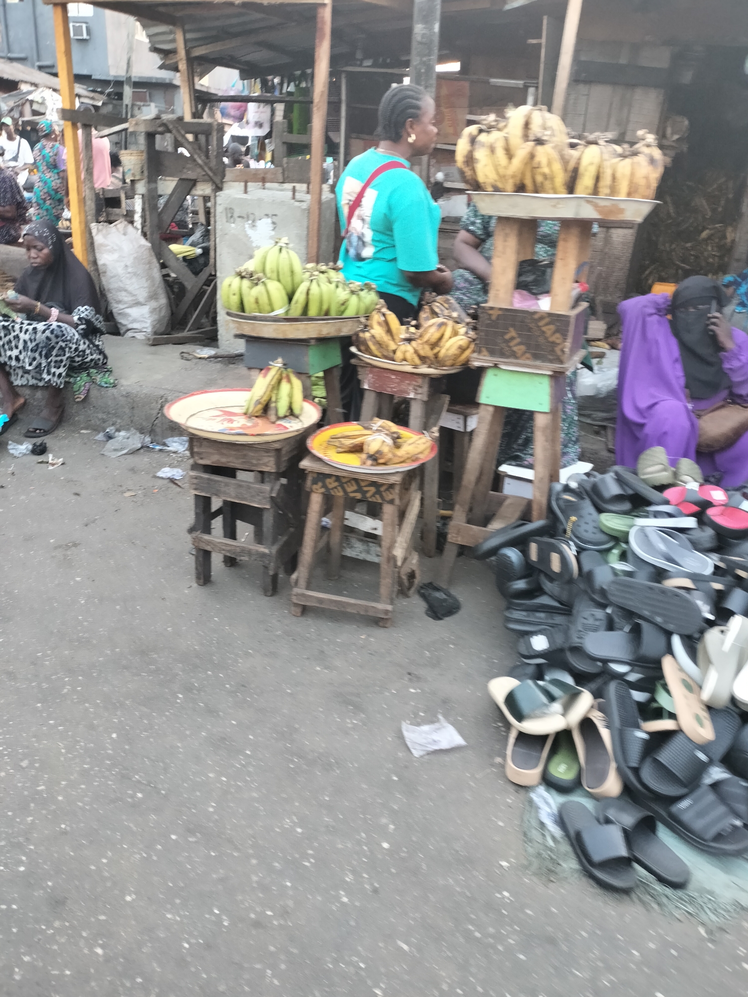 The image shows a street scene in Lagos, Nigeria, likely a marketplace. Several vendors are visible. One vendor wears a turquoise shirt and stands behind a table with ripe plantains. Another vendor, dressed in black and leopard print, sits on the ground to the left. A third vendor in purple attire sits on the right, talking on a phone. Bananas are displayed on stands of varying heights, and a pile of sandals and flip-flops is on the ground. The environment appears to be a busy, open-air market with makeshift stalls and various structures.