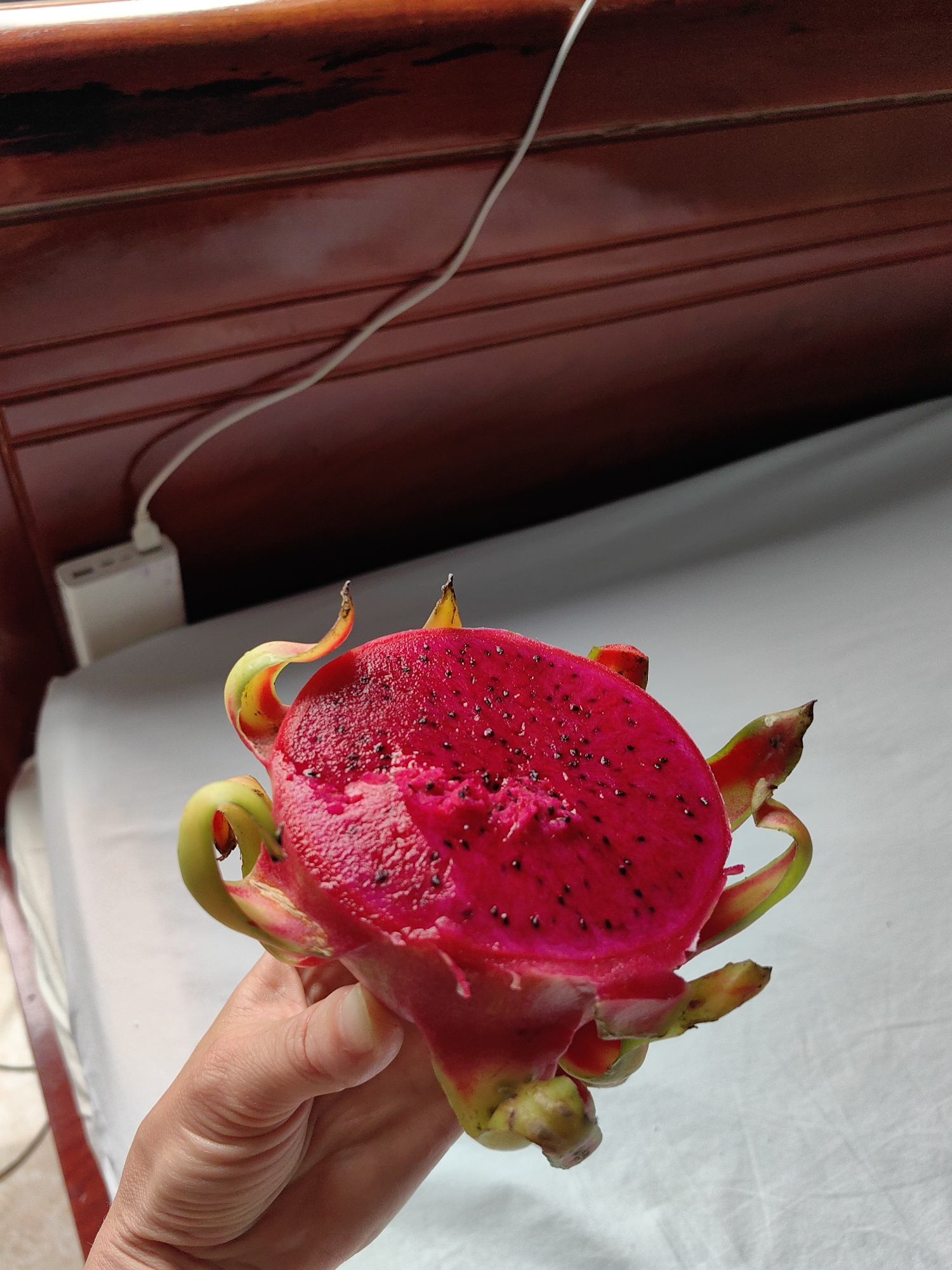 A hand holds a halved red dragon fruit in front of a bed in a room. The fruit's vibrant red flesh is speckled with small black seeds. The skin is pink with green and yellow tipped scales. The bed is covered in a white sheet. In the background is a varnished wood bed frame. A white cable connects to a white device near the head of the bed. No people are fully visible. The location is likely Bai Chay Ward, Vietnam.