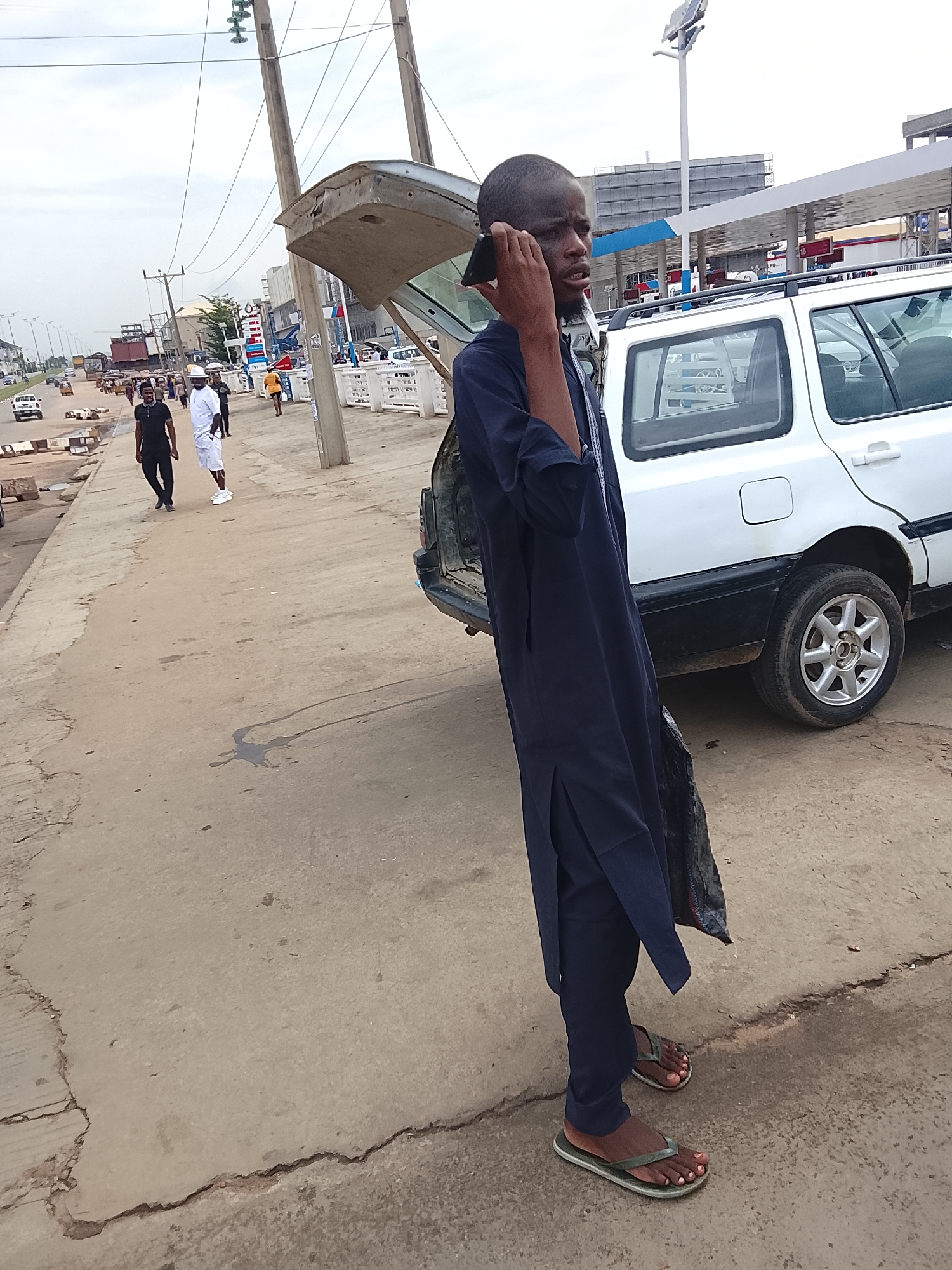 The image shows a man in Abuja, Nigeria, talking on his phone while standing next to a white car with its trunk open on a concrete sidewalk. The man is wearing a dark blue traditional outfit and sandals. In the background, there are other people walking on the sidewalk, a gas station, and buildings. Utility poles and overhead wires are visible. The weather appears cloudy. There is a visible oil-like stain on the pavement.