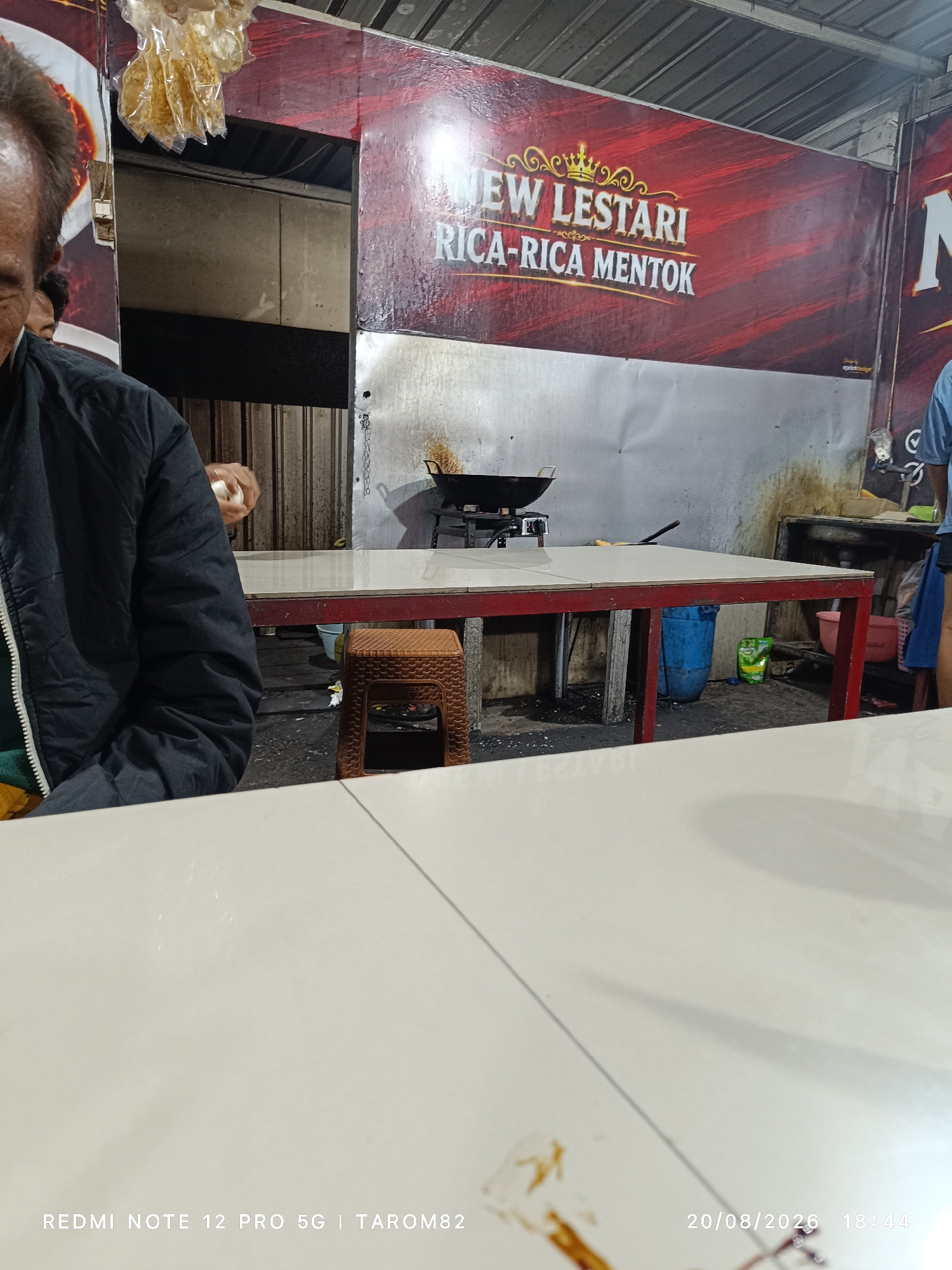This image captures a scene inside a food stall, likely a small, open-air establishment, in Bringinbendo, Indonesia. The background features a large red banner with gold lettering that reads "NEW LESTARI RICA-RICA MENTOK," indicating the name of the stall and a specialty dish. To the left, a portion of another banner shows a plate of food, possibly the signature dish. The stall appears to be set up for cooking and serving food, with a wok on a stove visible in the mid-ground.
In the foreground, a person, partially obscured, is seated at a white table, wearing a dark jacket. Another person, also partially visible, is standing near the cooking area. The environment is utilitarian, with stainless steel surfaces and a basic setup for a food business. The lighting suggests it is evening or indoors with artificial light. The overall impression is of a humble, functional eatery operating in a local Indonesian setting.