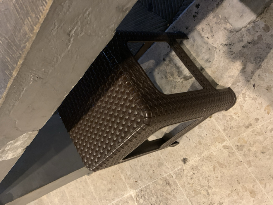 A brown plastic stool sits on a tiled floor in Probolinggo, Indonesia. The stool is made of woven plastic with a rectangular frame. The floor is made of square tiles with a slight beige color. There is a gray countertop partially visible in the upper portion of the image. The countertop is made of a smooth, hard material. The stool is positioned directly under the countertop. The scene appears to be in a bathroom, possibly in a residential home or public building. There is no evidence of people or activity in the image.