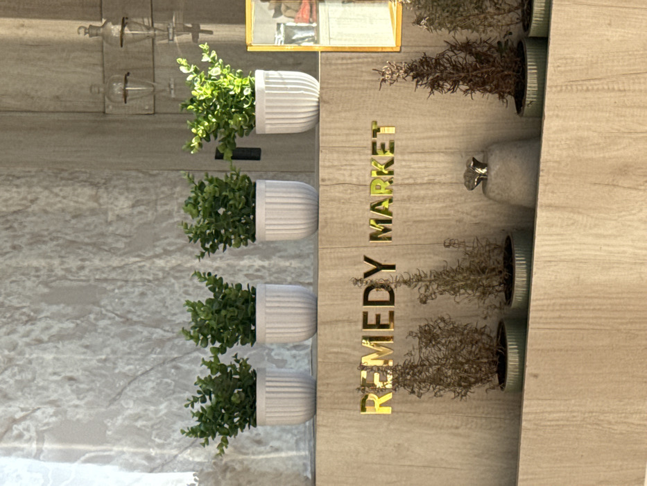 This image depicts an indoor scene of a retail space, identified as "REMEDY MARKET," located in Puebla City, Mexico. An upper horizontal wooden shelf displays four evenly spaced white, ribbed cylindrical pots, each containing a small, green artificial plant. Below this shelf, on a vertical wooden panel with a light wood grain finish, gold-colored, reflective block letters spell "REMEDY MARKET." A lower horizontal wooden shelf is situated beneath the signage, holding five light green, ribbed cylindrical pots containing dried or artificial brown-hued plants. Between the third and fourth lower pots from the left, a light gray, bell-shaped object with a metallic silver finial rests. The background wall features a light gray, mottled pattern. To the right of the upper shelf, two clear glass vessels with inverted stoppers are wall-mounted. A partial view of a gold-framed rectangular object, possibly a mirror or display case, is visible on the far right, reflecting indistinct red and white items. Illumination originates from the right, casting soft shadows and highlighting elements. No individuals are present.