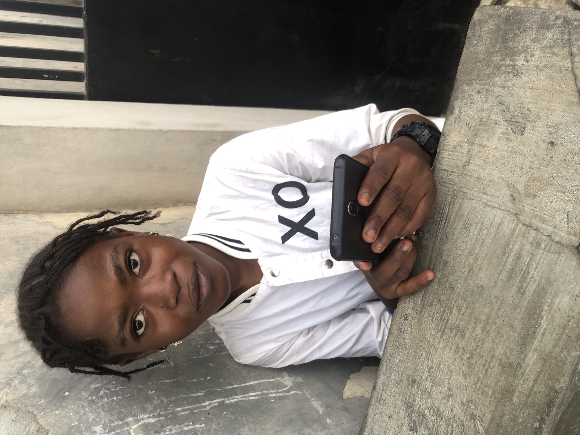 A young person with dark skin and braided hair is leaning against a concrete structure, looking towards the camera. They are wearing a white shirt with black trim on the collar and cuffs, and a black graphic that appears to be "XO" on the chest. The individual is holding a black smartphone in their hands. The background consists of a textured gray wall on the left and a section of a building with vertical black bars and a black door on the right. The lighting suggests it is daytime. The scene appears to be outdoors, possibly in a residential or urban area of Ado, Nigeria.
