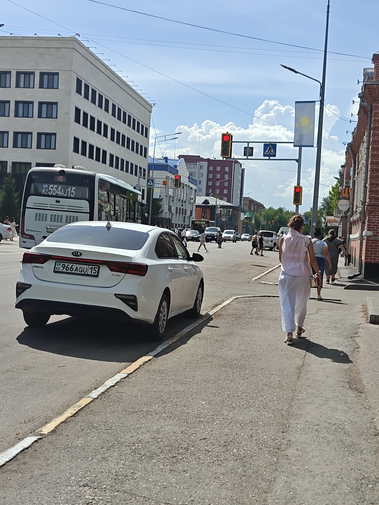The image depicts a street scene in Petropavl, Kazakhstan, on a sunny day with a partly cloudy sky. In the foreground, a white Kia sedan with license plate "KZ 966 AGU 15" is parked on the right side of the road. The car is facing away from the camera. A woman wearing a light pink shirt and white pants is walking away from the camera on the sidewalk to the right of the car. Further down the street, a white bus with the number "16" and license plate "KZ 554 AJ 15" is visible, along with other vehicles and pedestrians. Several buildings line the street, including a modern, light-colored building on the left and a red brick building on the right. Traffic lights and street signs are also present, indicating an urban environment. The road has distinct yellow and white markings.
